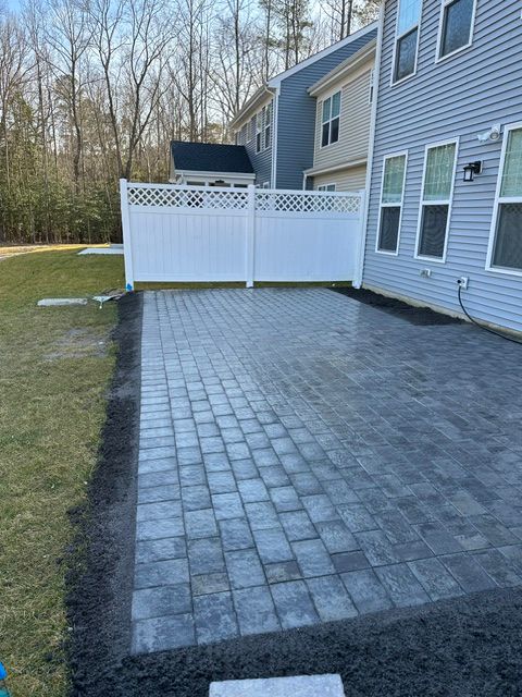 Paver patio beside a blue house with white lattice fence, bordered by grass and trees.