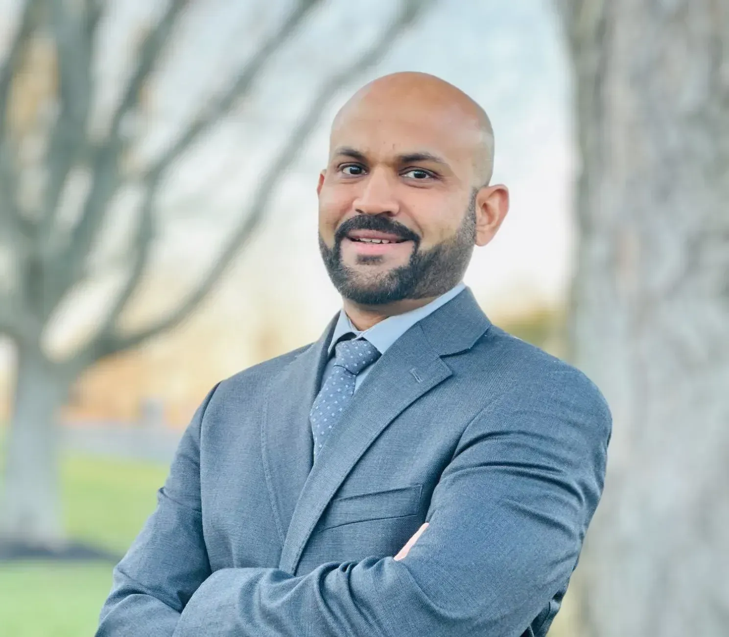 A man in a suit and tie is standing with his arms crossed in front of a tree.