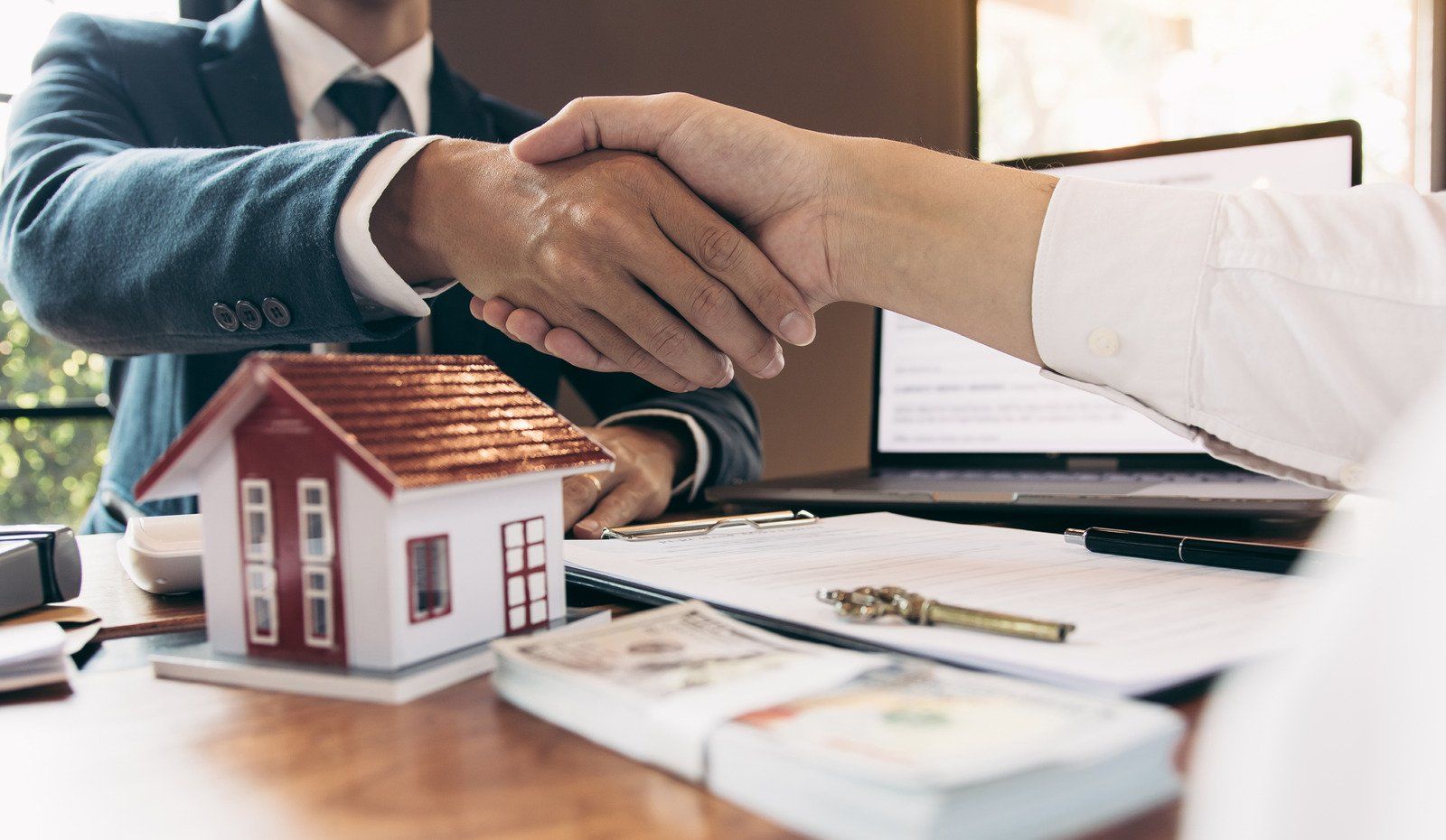 People shaking hands over a desk with a house model, documents, and cash, symbolizing a real estate deal.