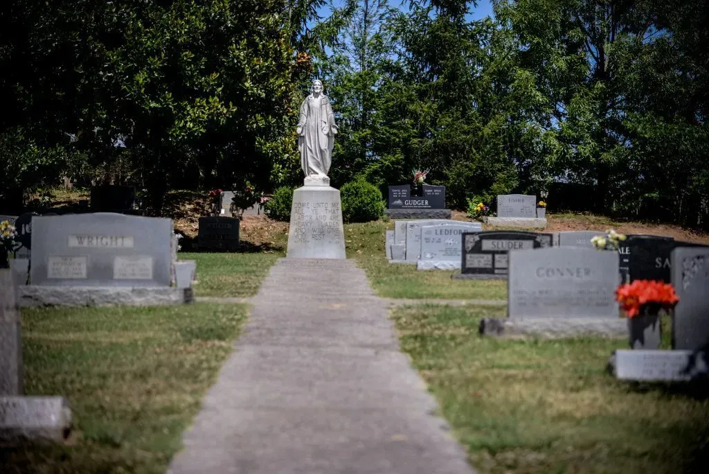 Cemetery with stone paths, headstones, and a statue of a robed figure on a pedestal under a blue sky.