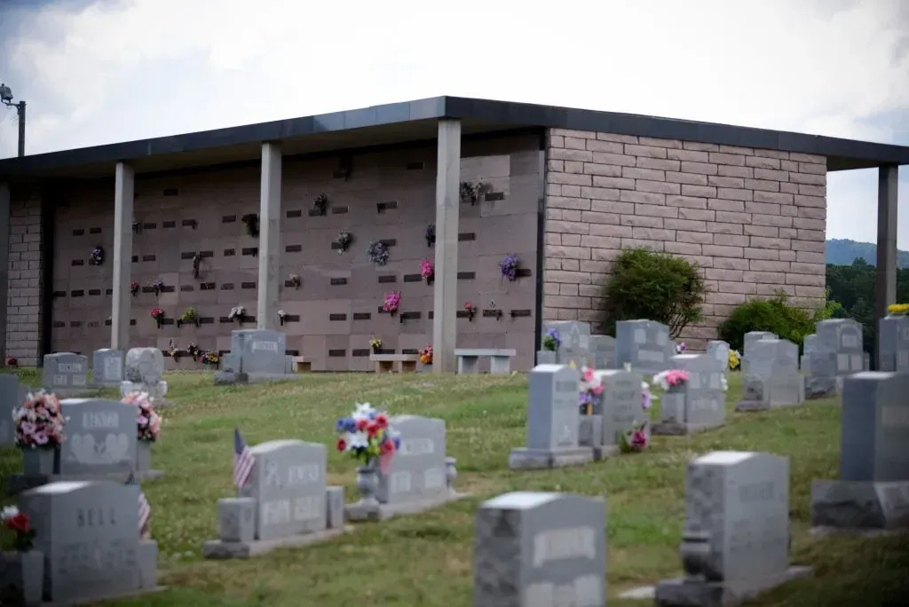 Cemetery scene with headstones in the foreground, a mausoleum with niches and columns, and a cloudy sky.