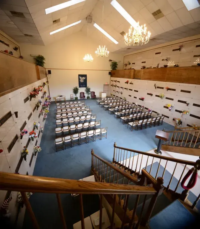Interior of a mausoleum, rows of chairs face a podium, flower-filled crypts line walls, chandeliers.