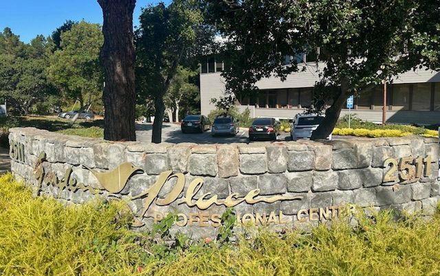 Stone sign for “The Place” at Rose Medical Center with trees and parked cars in the background