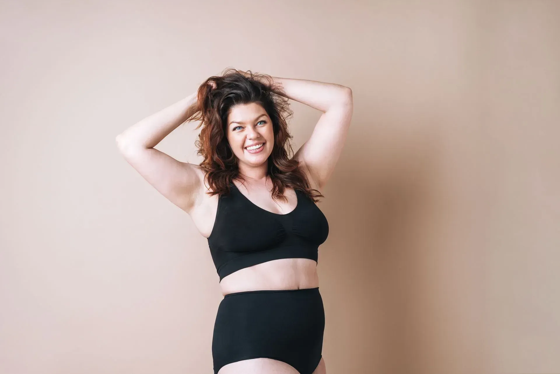 Woman in black sportswear posing with hands behind her head against a beige wall