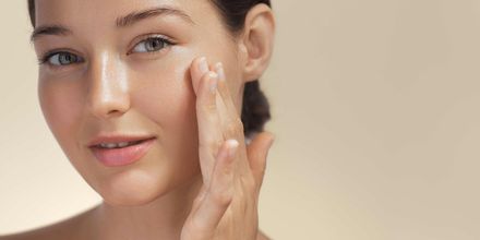 Close-up of a woman applying facial cream to her cheek against a soft beige background