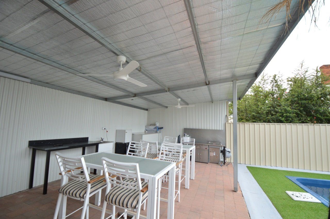 Covered outdoor dining area with tables, chairs, and a pool. White walls, a metal roof, and green turf are visible.