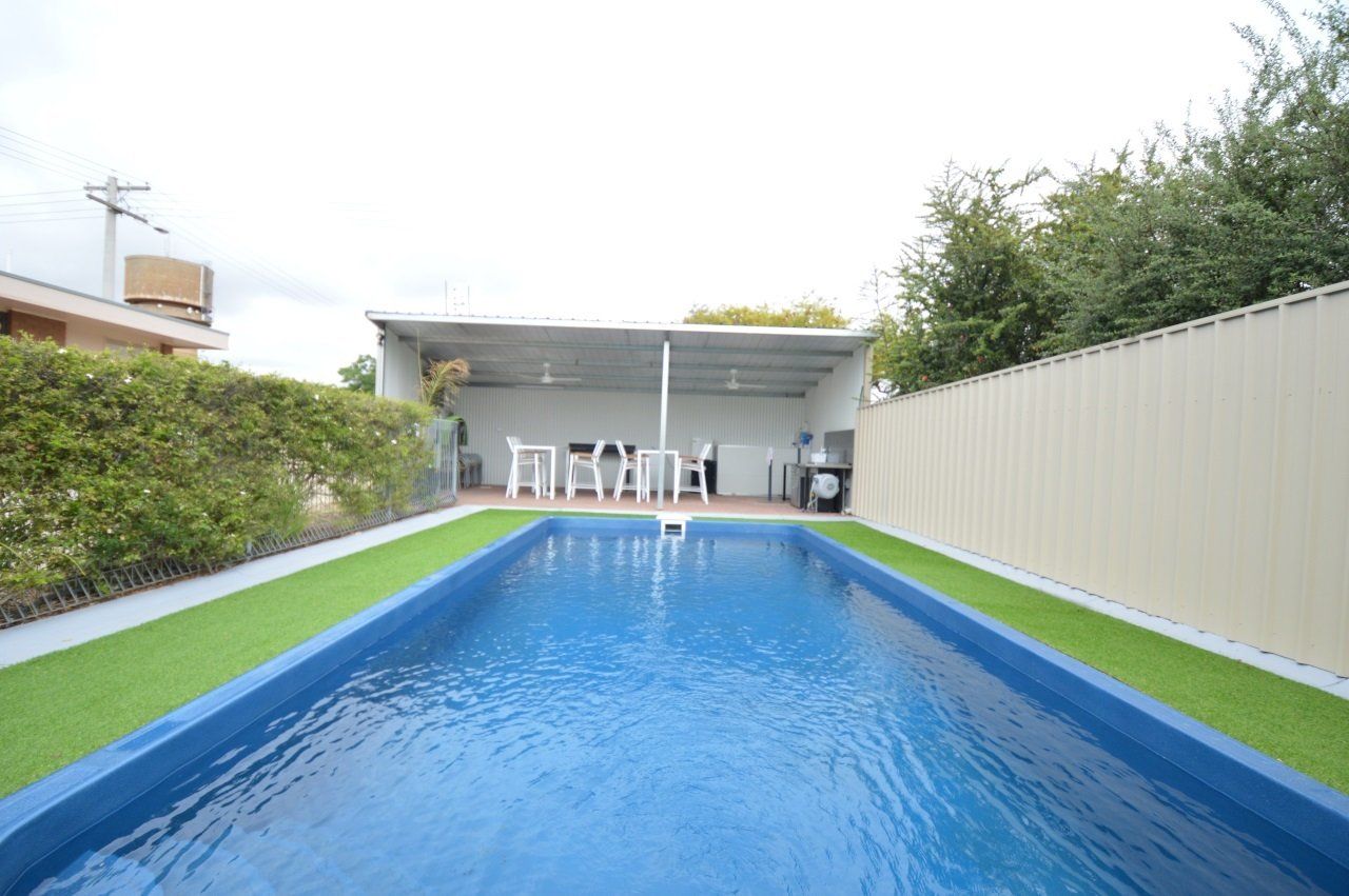 Long, blue swimming pool with surrounding artificial turf and a covered patio area with seating.