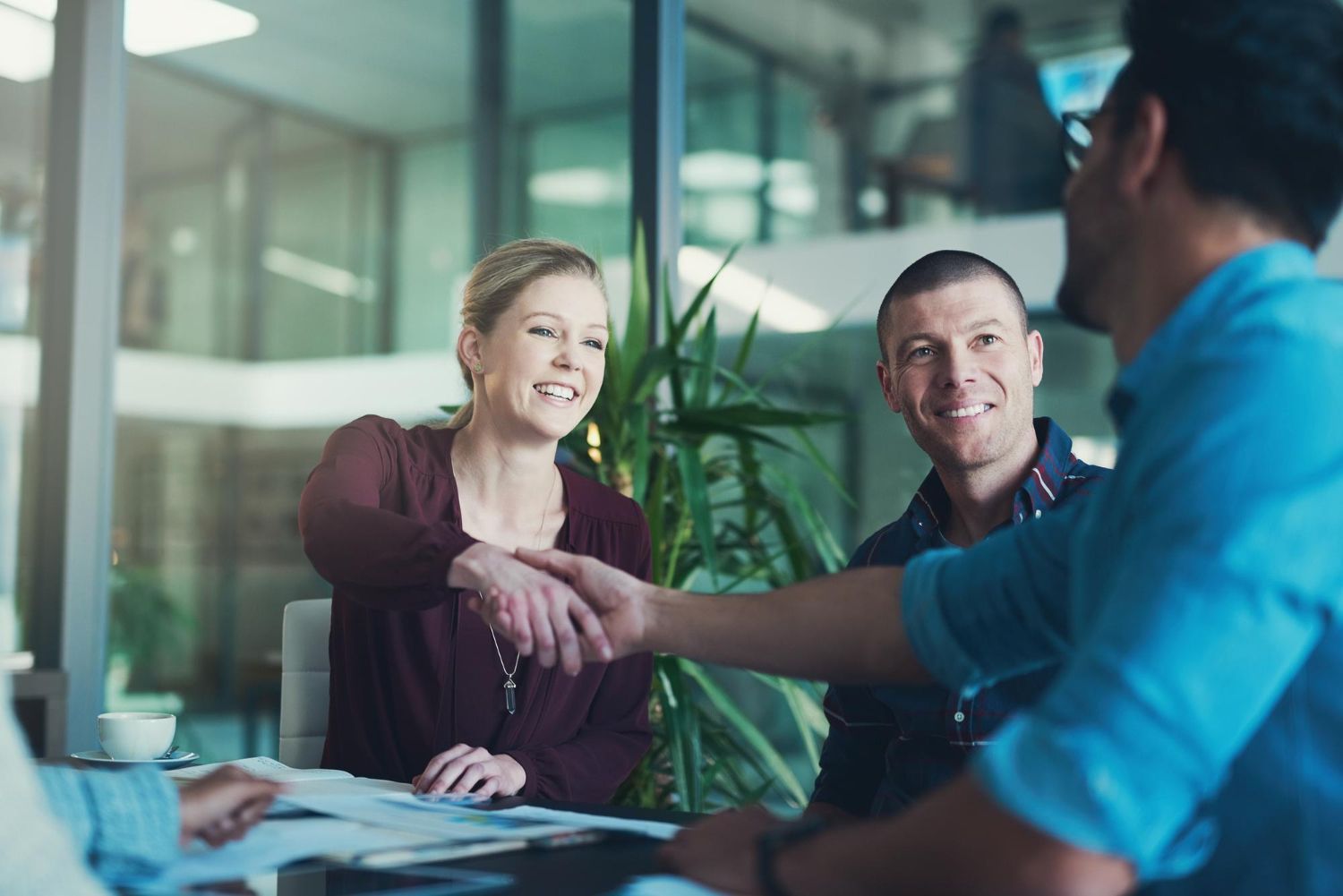 People shaking hands in an office, smiling. Meeting at a table with papers, natural light, and glass wall.