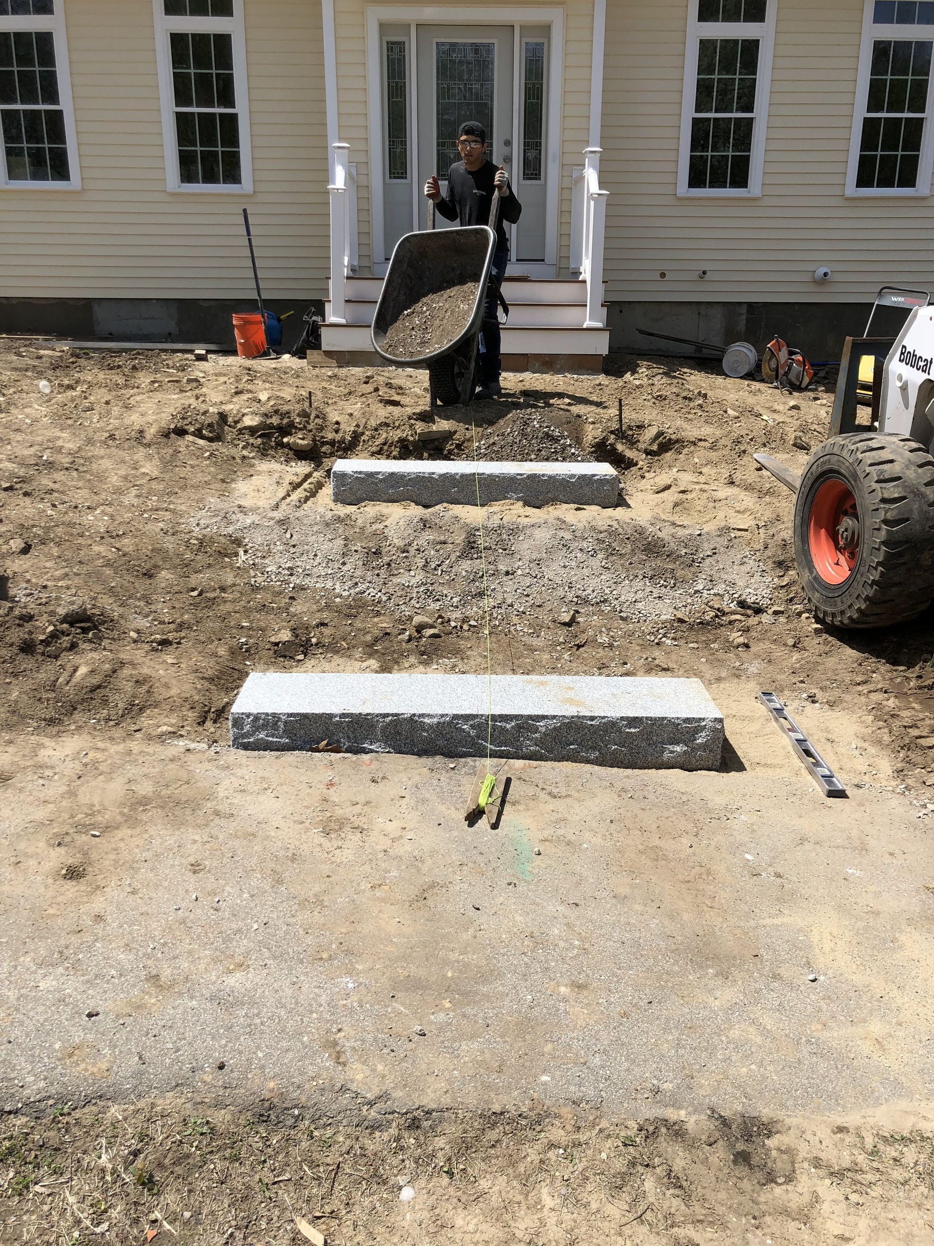 Construction of steps leading to a house. A worker dumps gravel. Bobcat on the right.