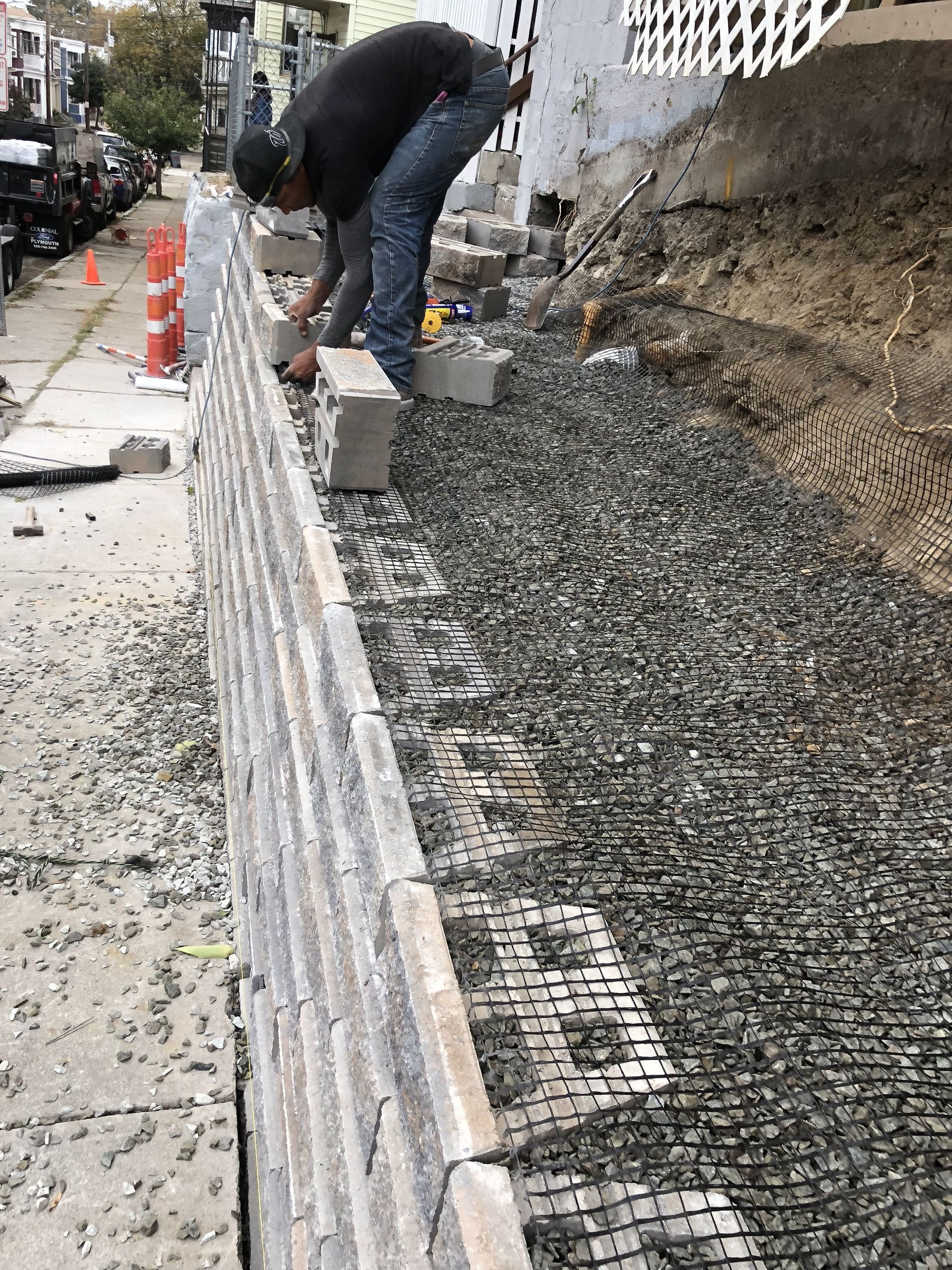 Man building a retaining wall on a city sidewalk with gray bricks and gravel.