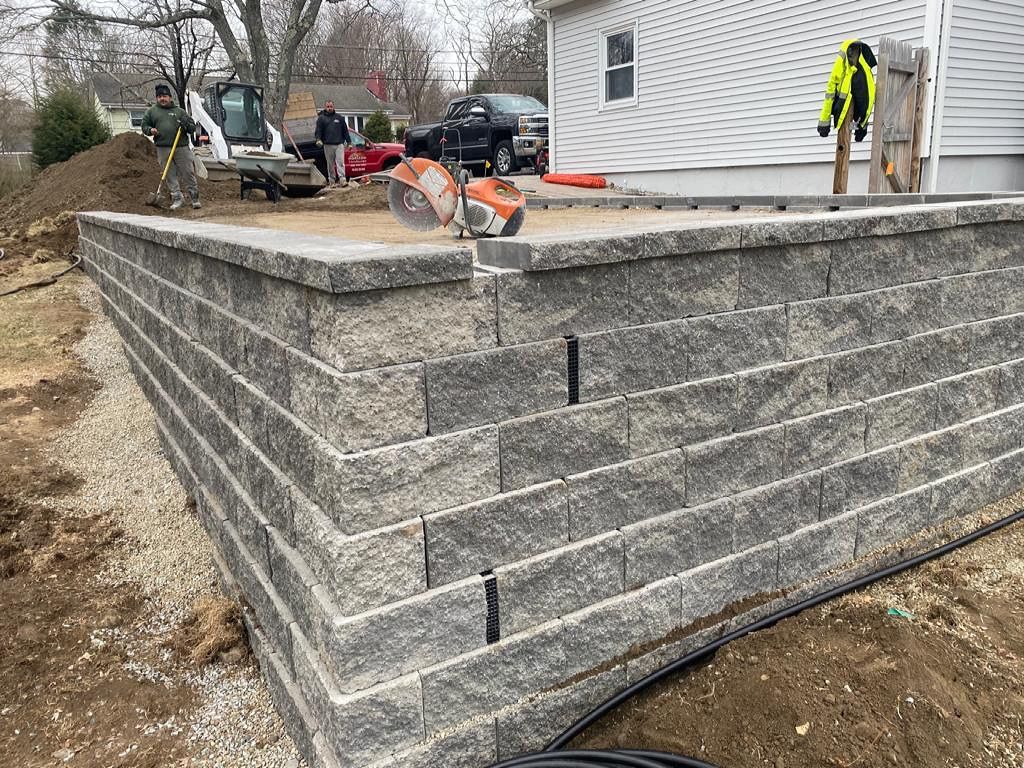 Retaining wall construction using gray interlocking blocks; tools and workers are visible in the background.