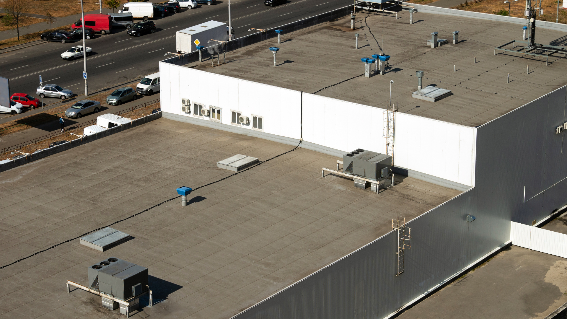 Aerial view of a large flat-roof industrial building beside a highway, with rooftop equipment and parked vehicles.
