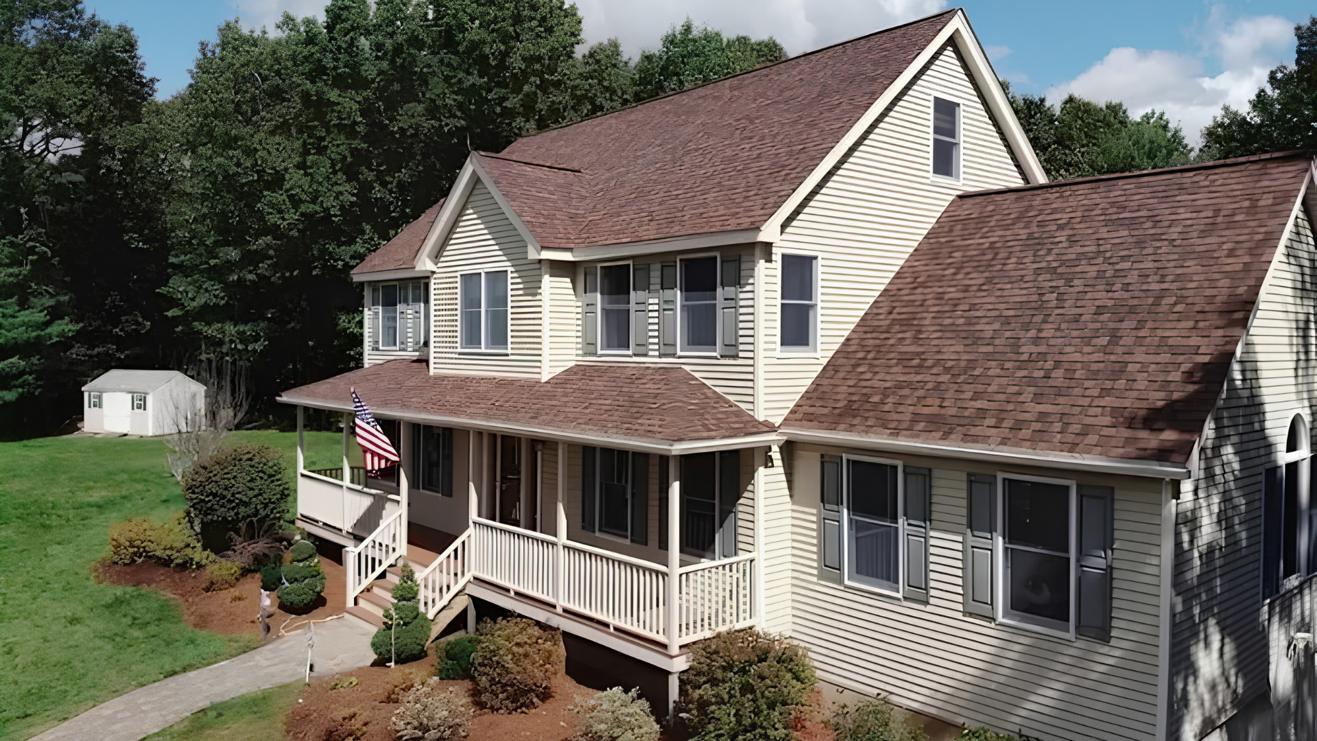 Two-story beige house with brown roof and covered porch, surrounded by trees and lawn