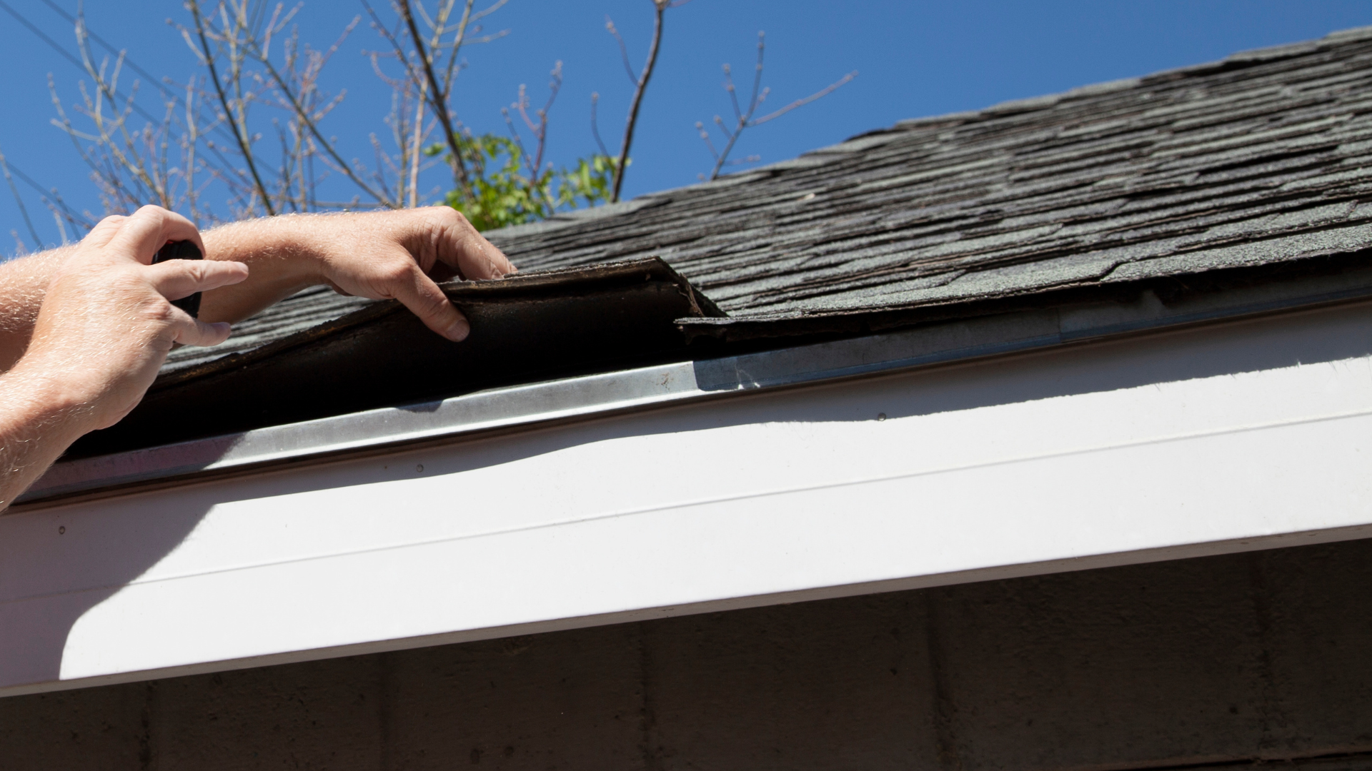 Hands inspecting and lifting roof shingles at the edge of a house roofline