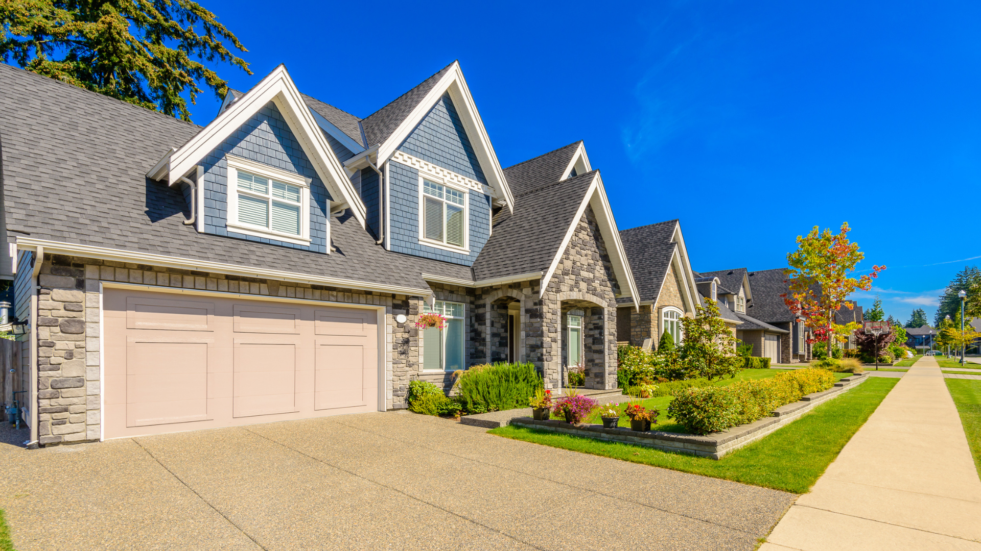 Suburban house with gray siding, beige garage, stone entry, and manicured lawn along a sidewalk