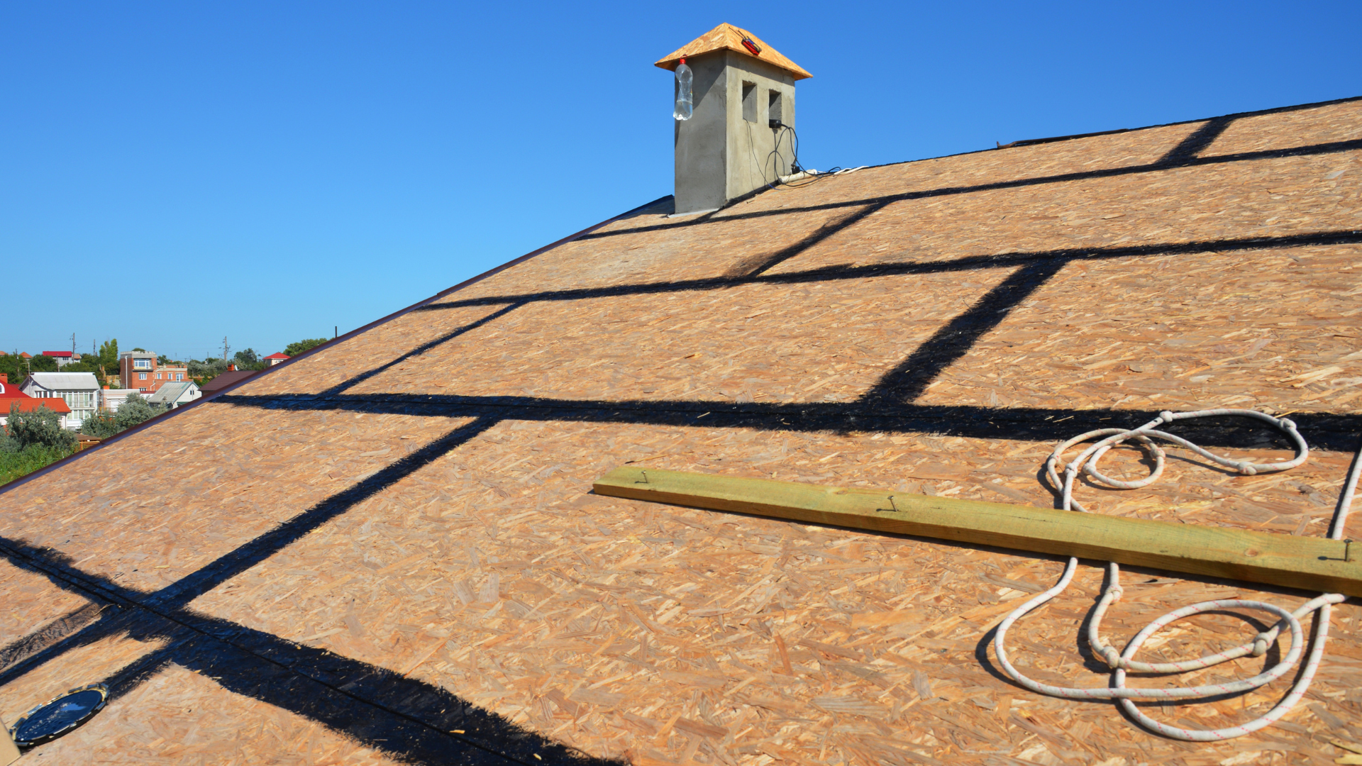 Roof under construction with brown shingles, wooden boards, and coiled rope against a blue sky
