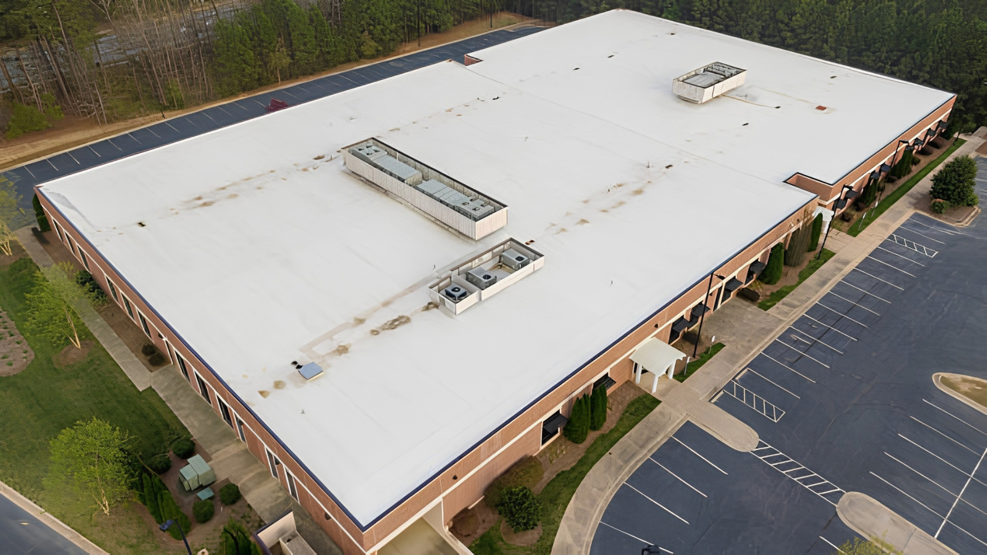 Aerial view of a large white-roofed commercial building with parking lot and surrounding trees