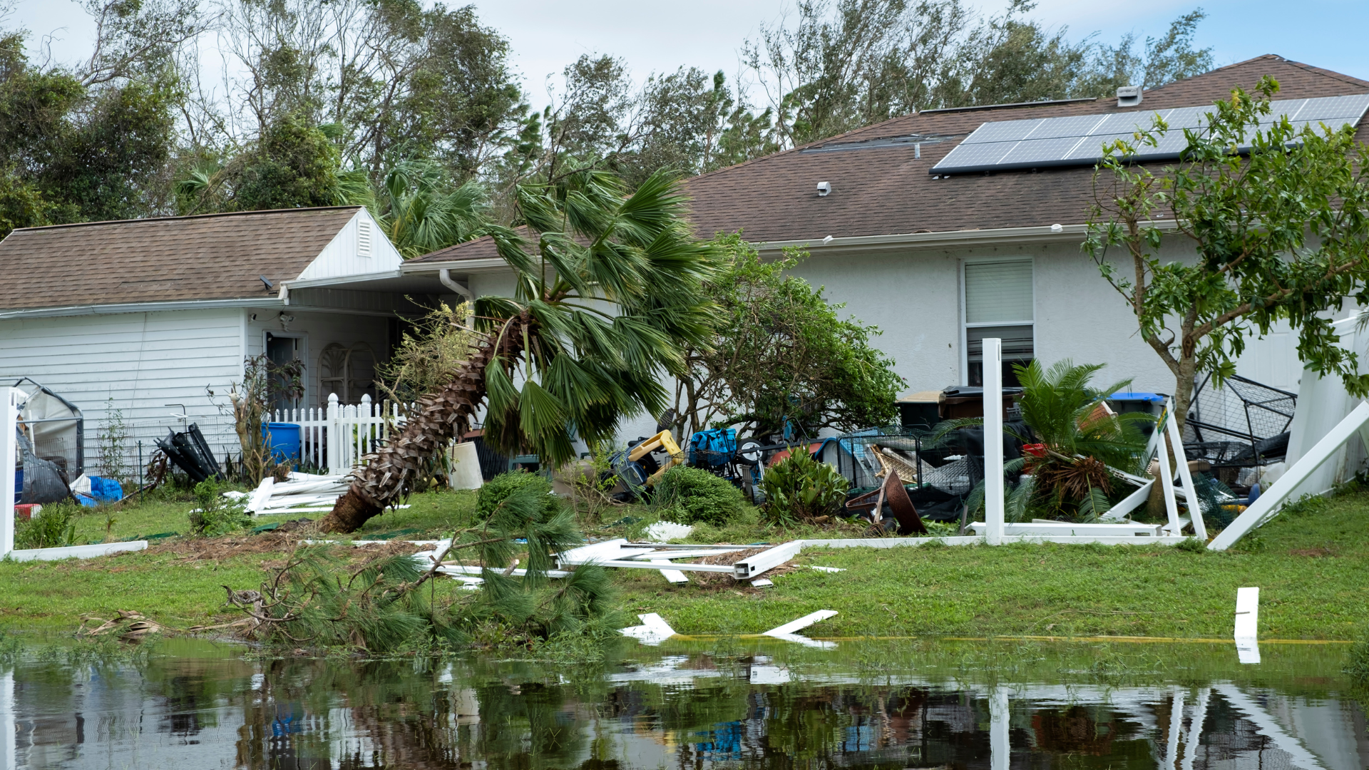 Damaged homes with debris and broken trees after storm flooding, reflected in standing water