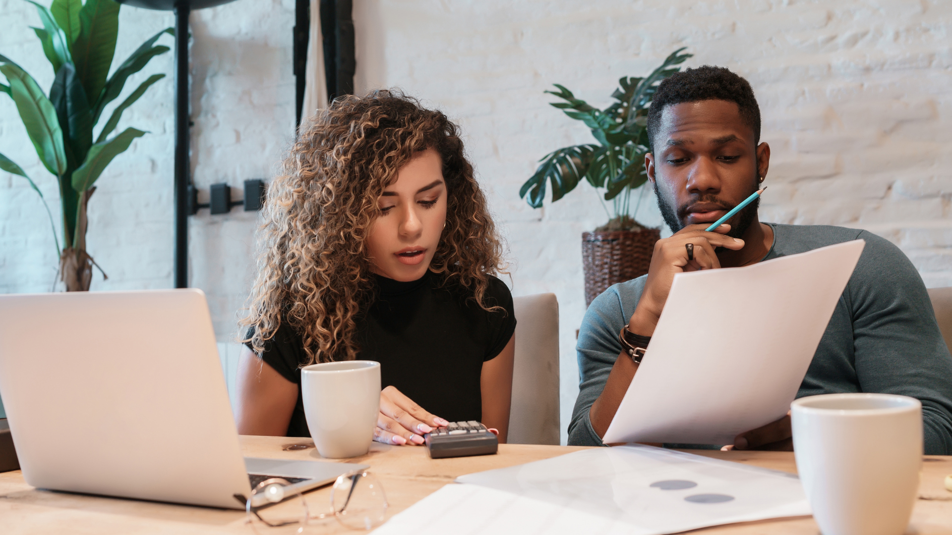 Two people working at a table with laptops, papers, and coffee cups in a bright office.