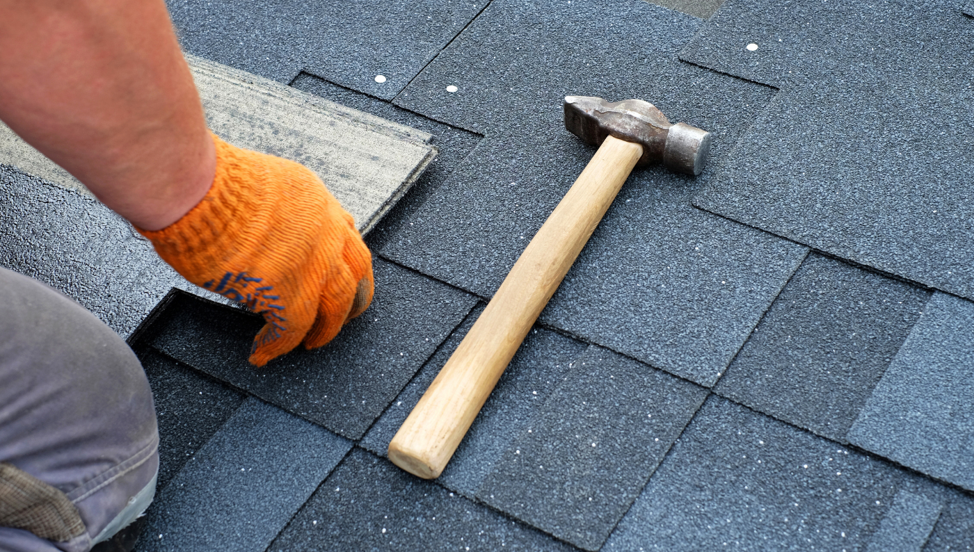 Gloved hand lifting asphalt shingles beside a hammer on a roof.