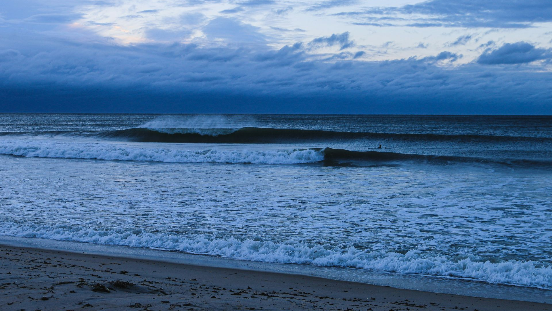 Blue ocean waves under a cloudy sky at dusk, washing onto a dark sandy beach.