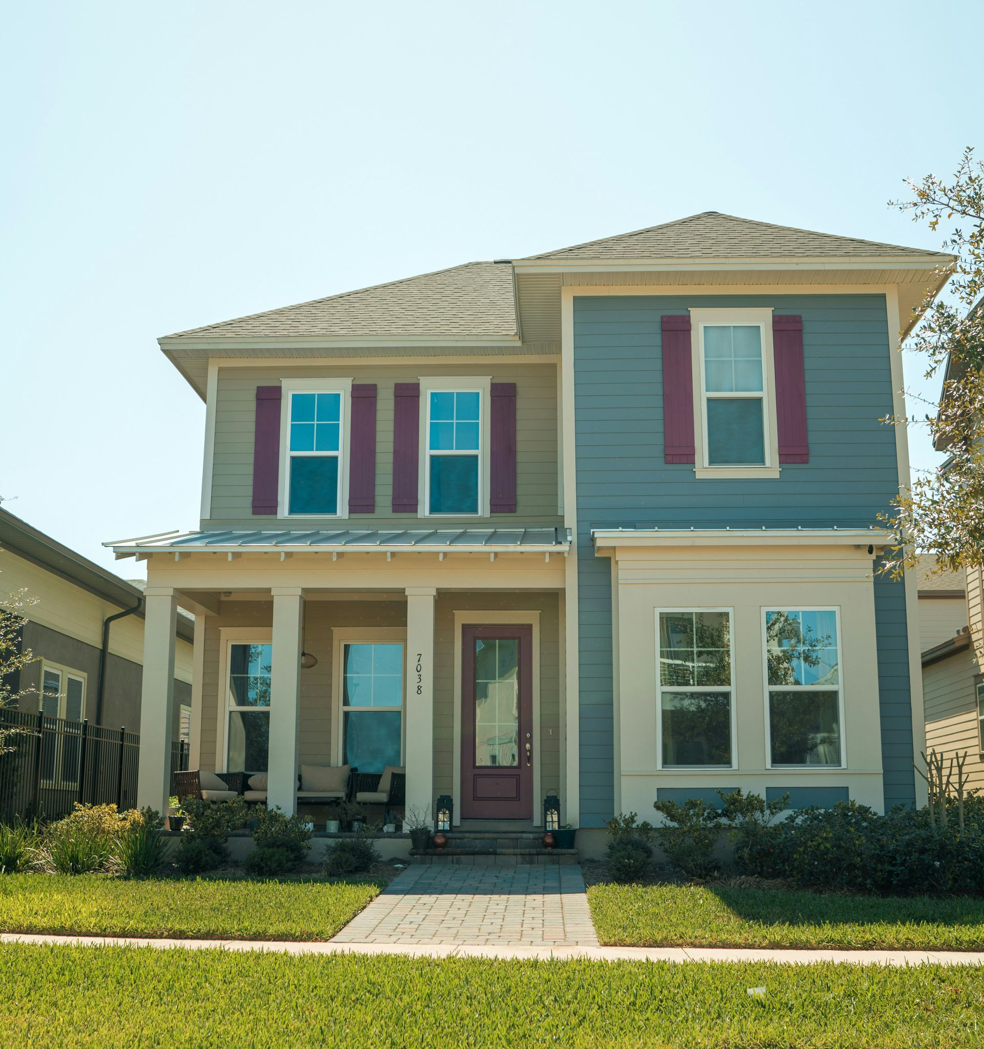 Two-story blue house with purple shutters, front porch, and small front yard in a sunny neighborhood.