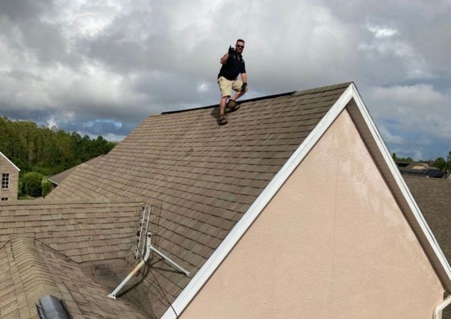 Person kneeling on a shingled roof beside a beige house under cloudy skies