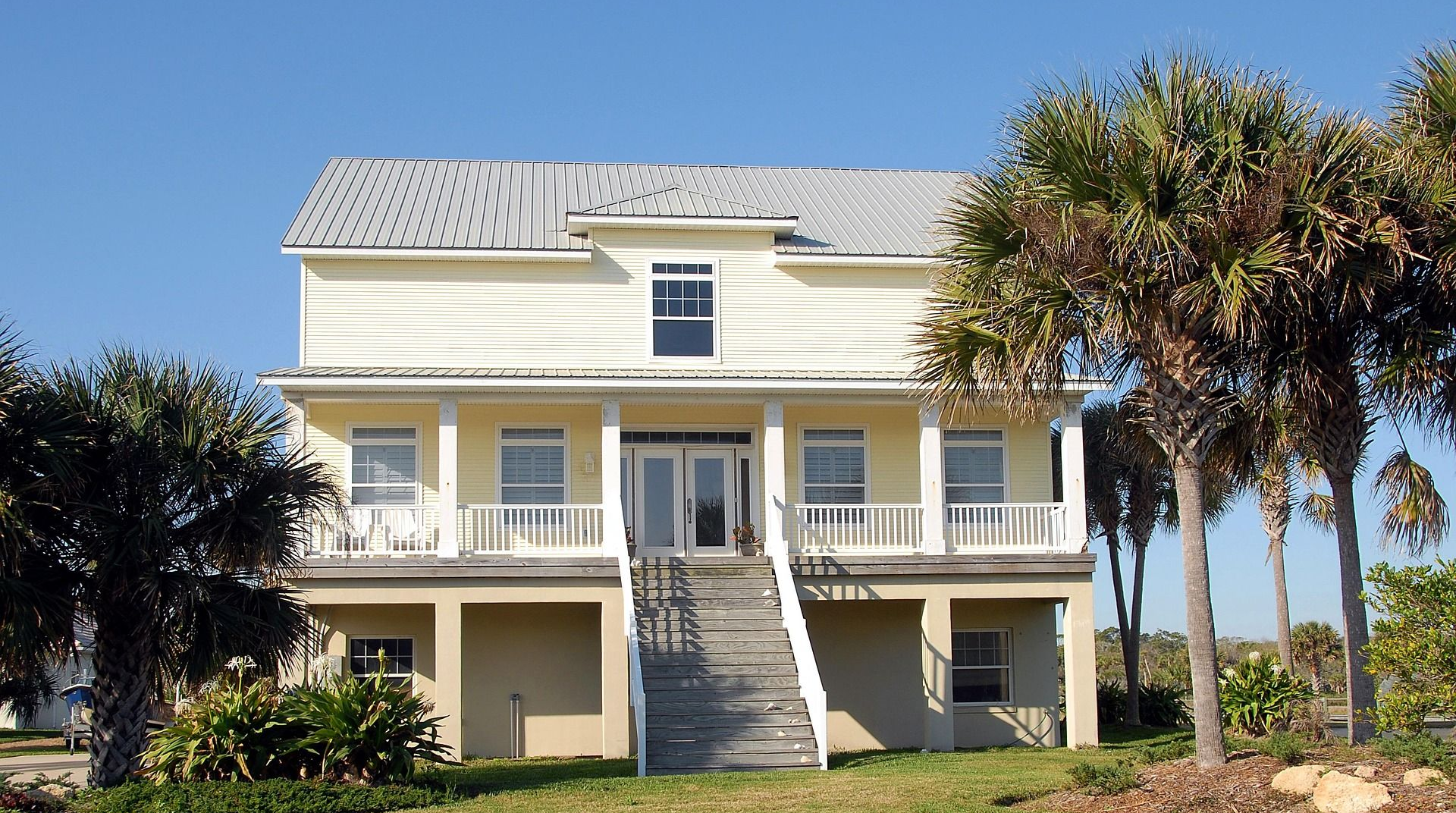 Two-story beach house with white siding, central stairs, and palm trees under a clear blue sky