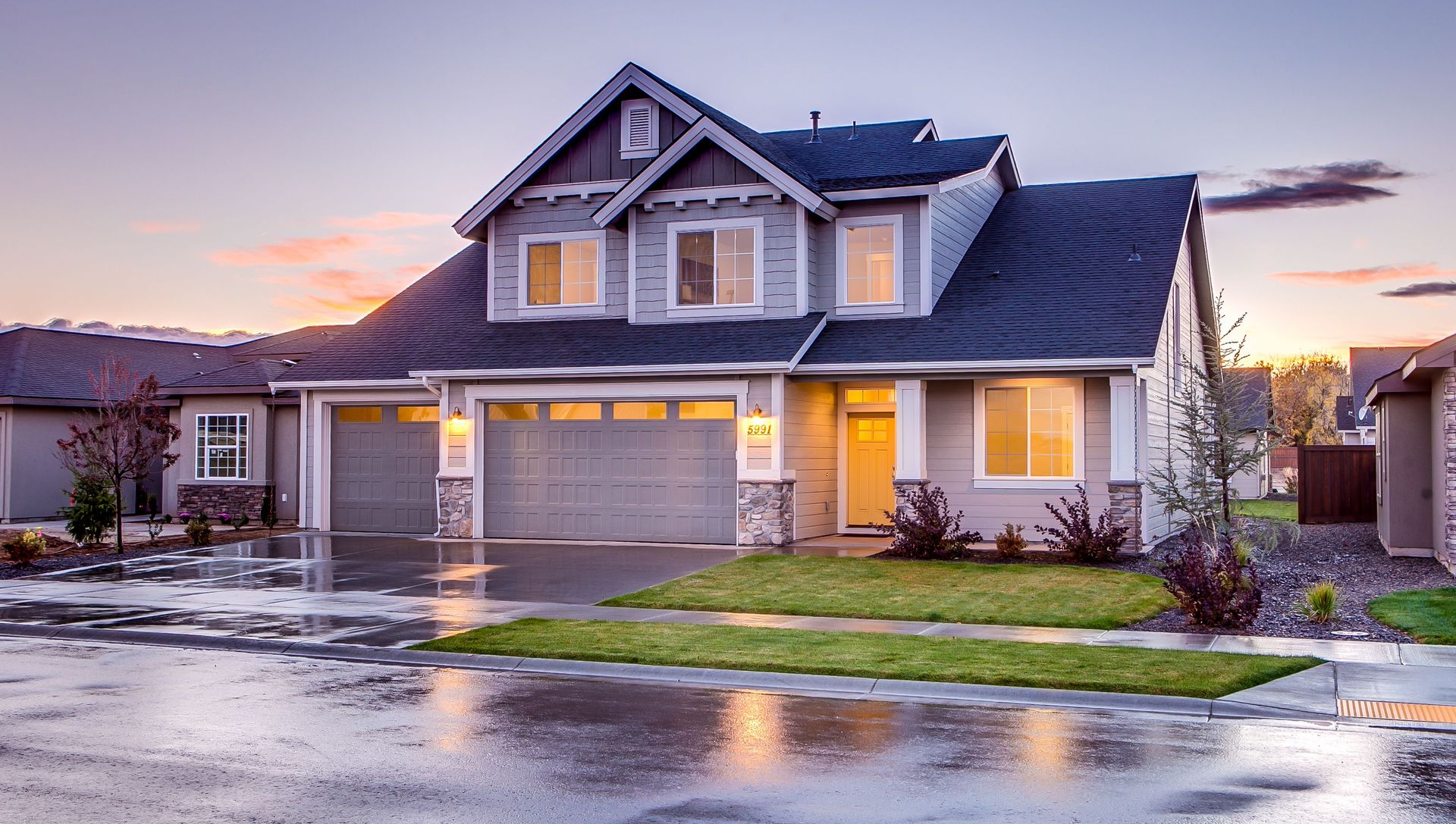 Suburban house at dusk with warm lights reflecting on wet pavement, under a cloudy sky