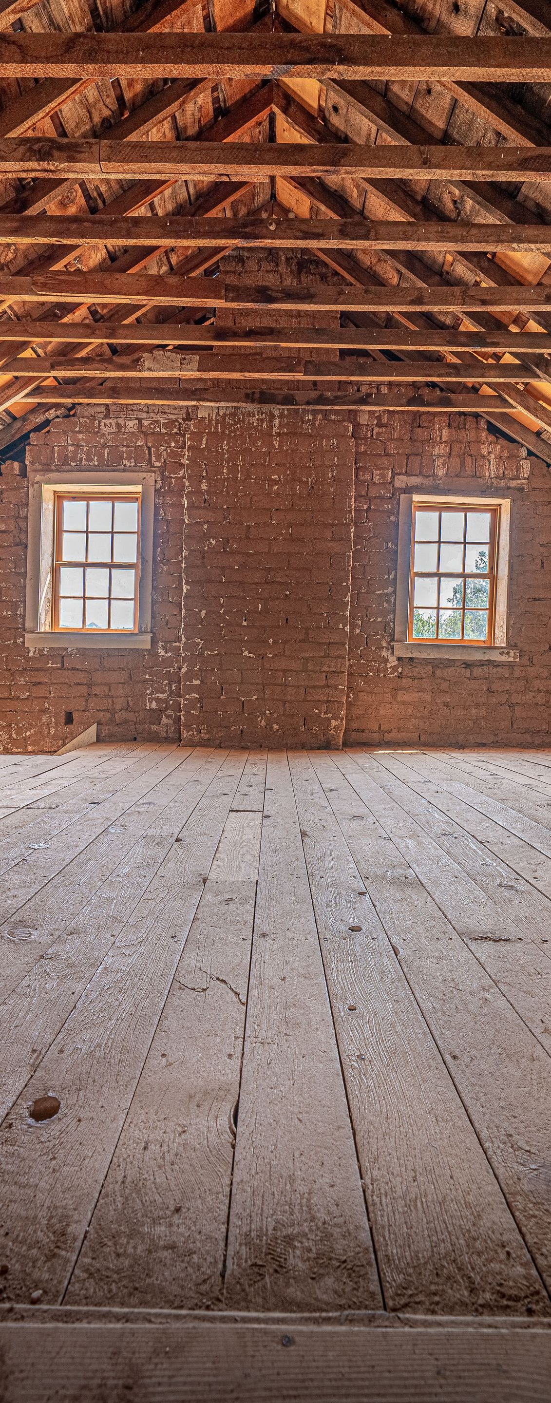 Empty rustic room with exposed wood ceiling, earthen walls, two windows, and a worn wooden floor