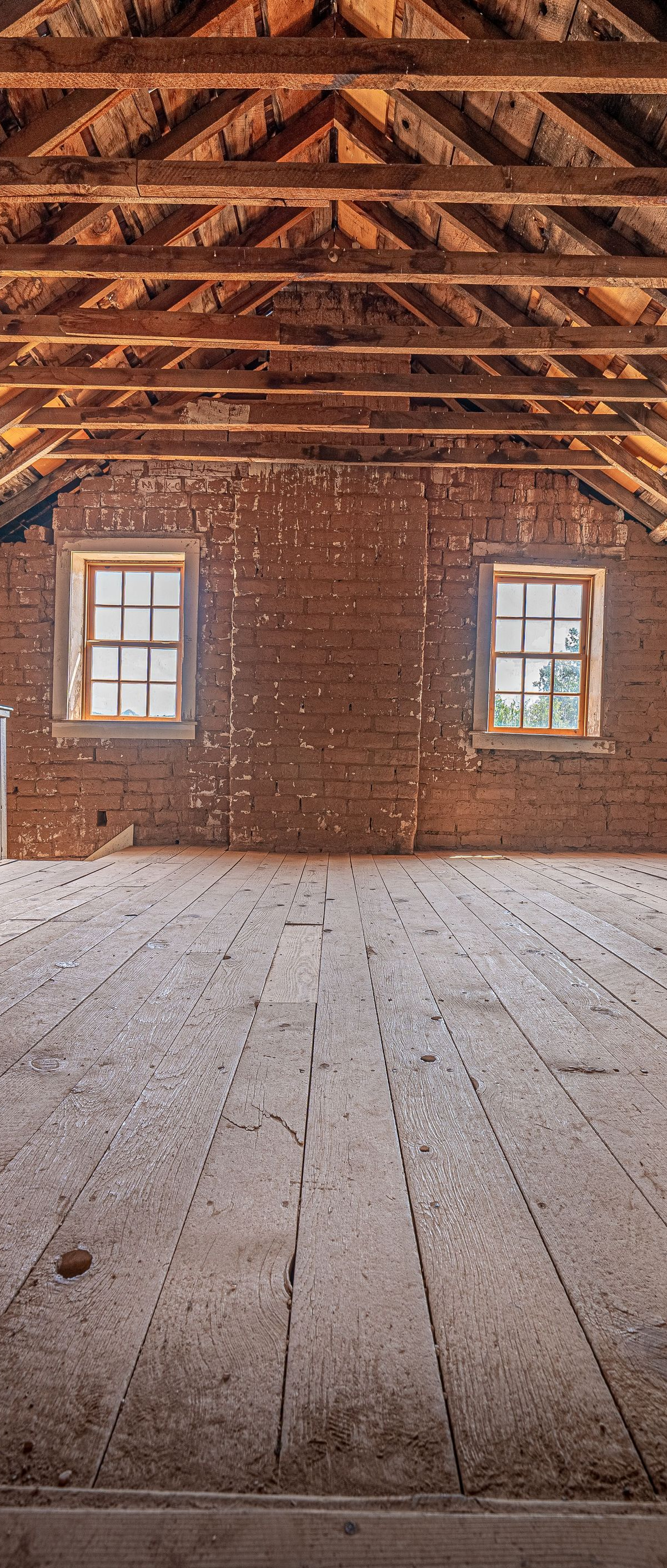 Empty rustic attic with exposed wooden beams, two small windows, and weathered plank floor
