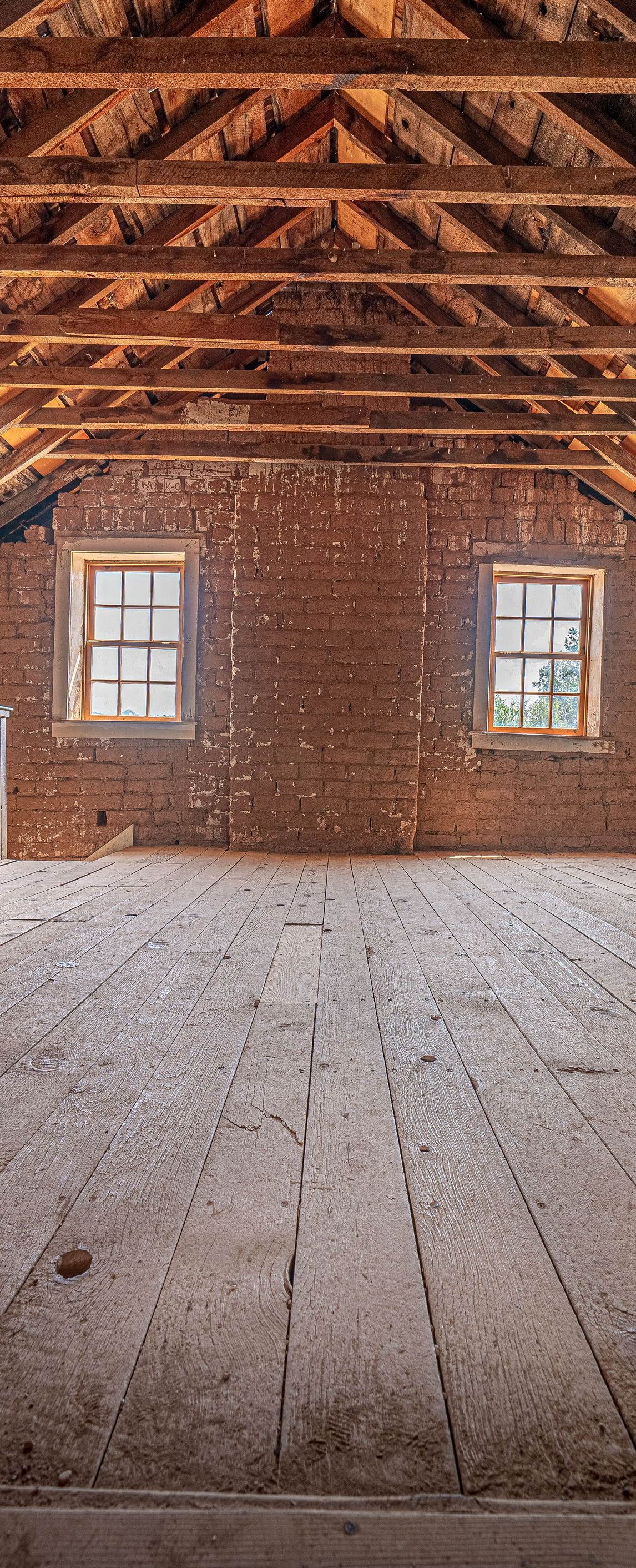 Empty rustic attic with exposed wooden beams, two small windows, and weathered plank floor