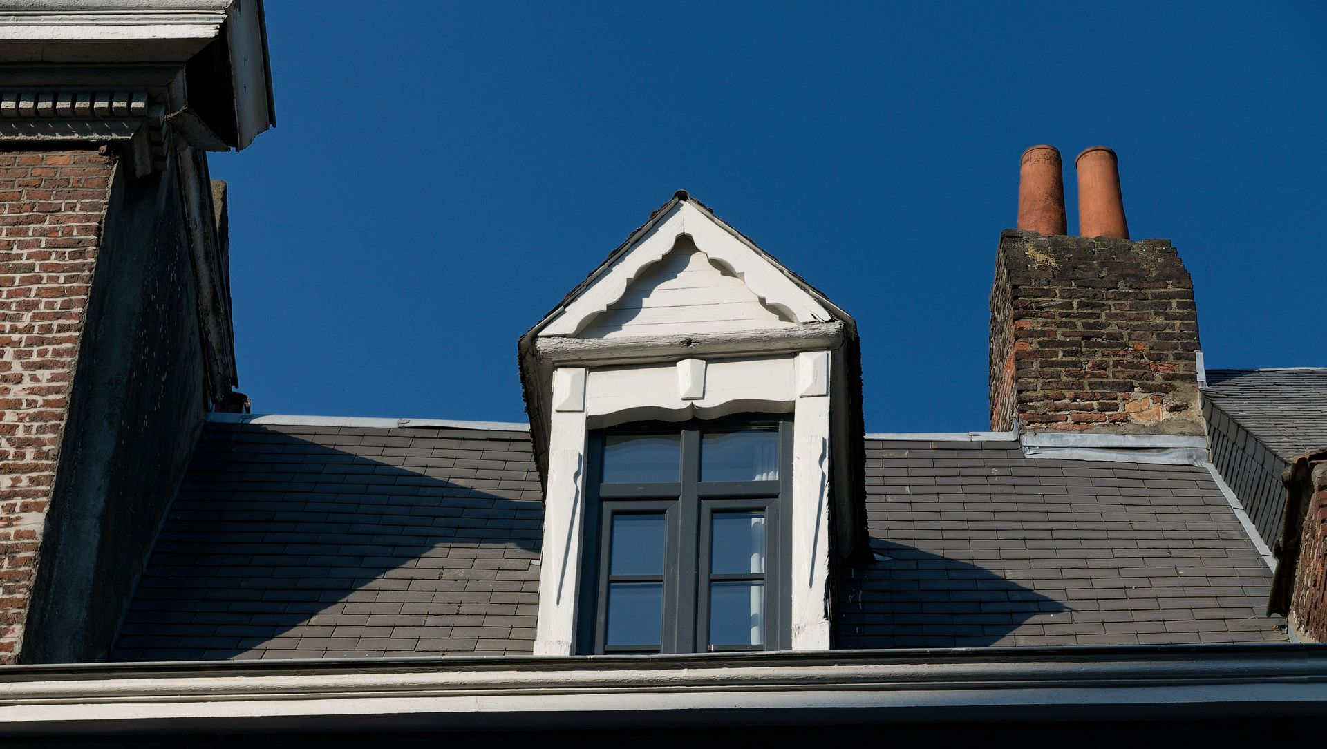 Dormer window on a gray slate roof against a blue sky, with brick chimneys on either side
