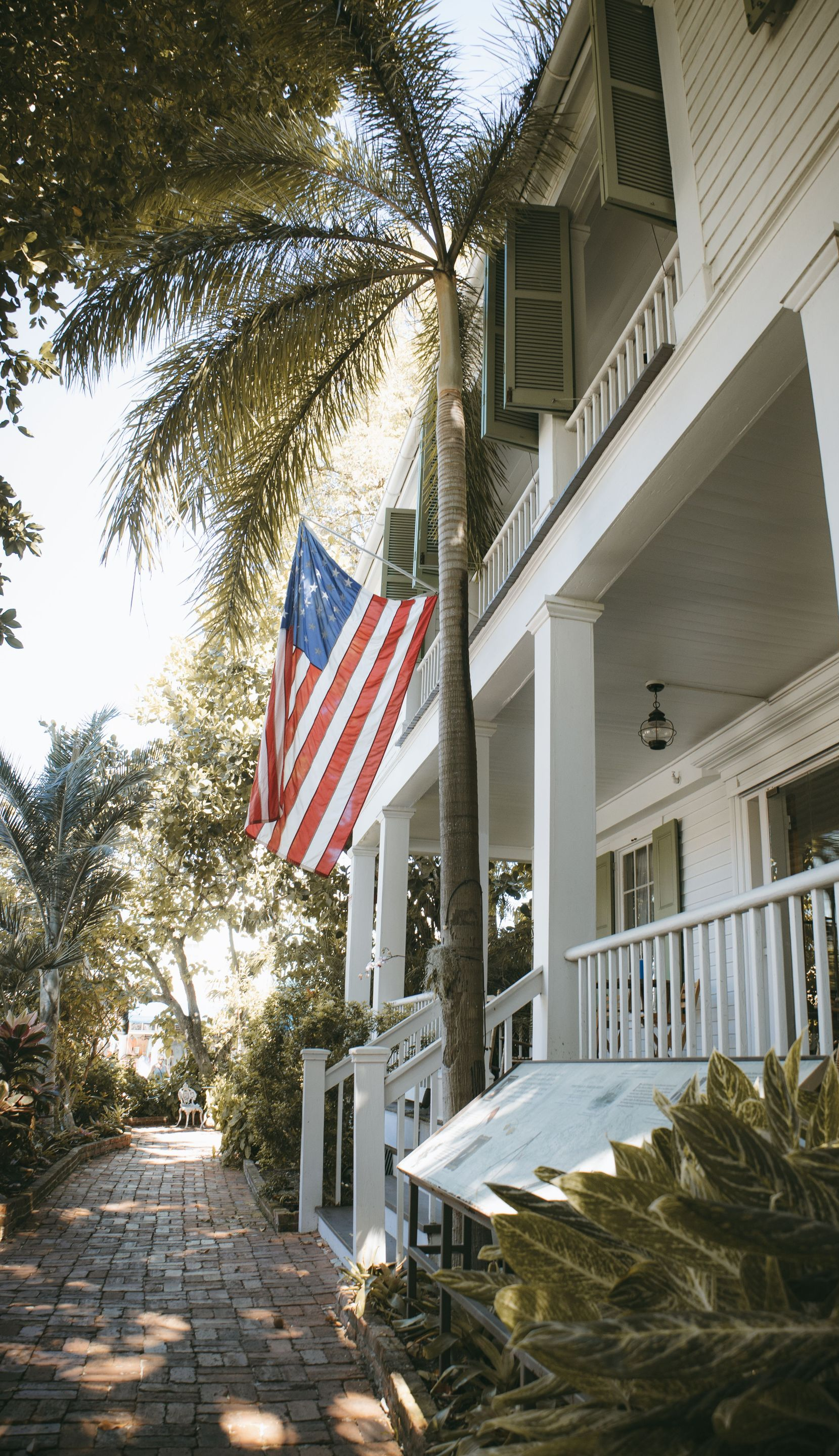 American flag hanging outside a white house with porch, shaded by palm trees along a brick walkway