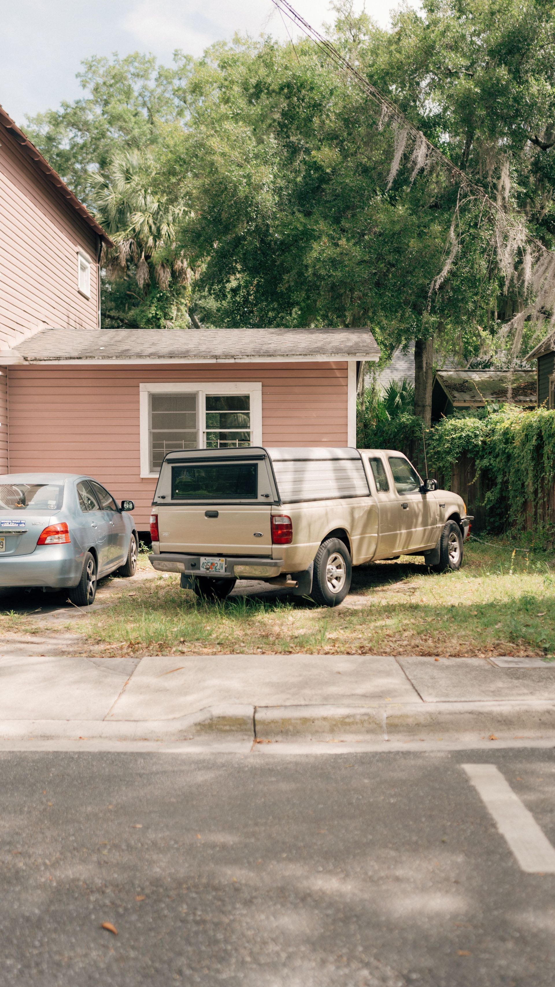 Parked silver sedan and beige pickup in front of a pink house with trees and driveway