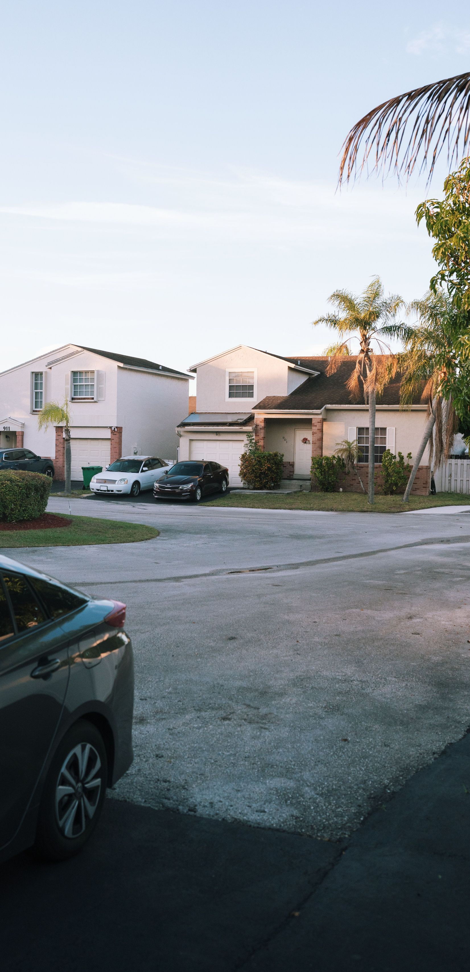 Residential parking lot with two white houses, a parked car, and palm trees at dusk