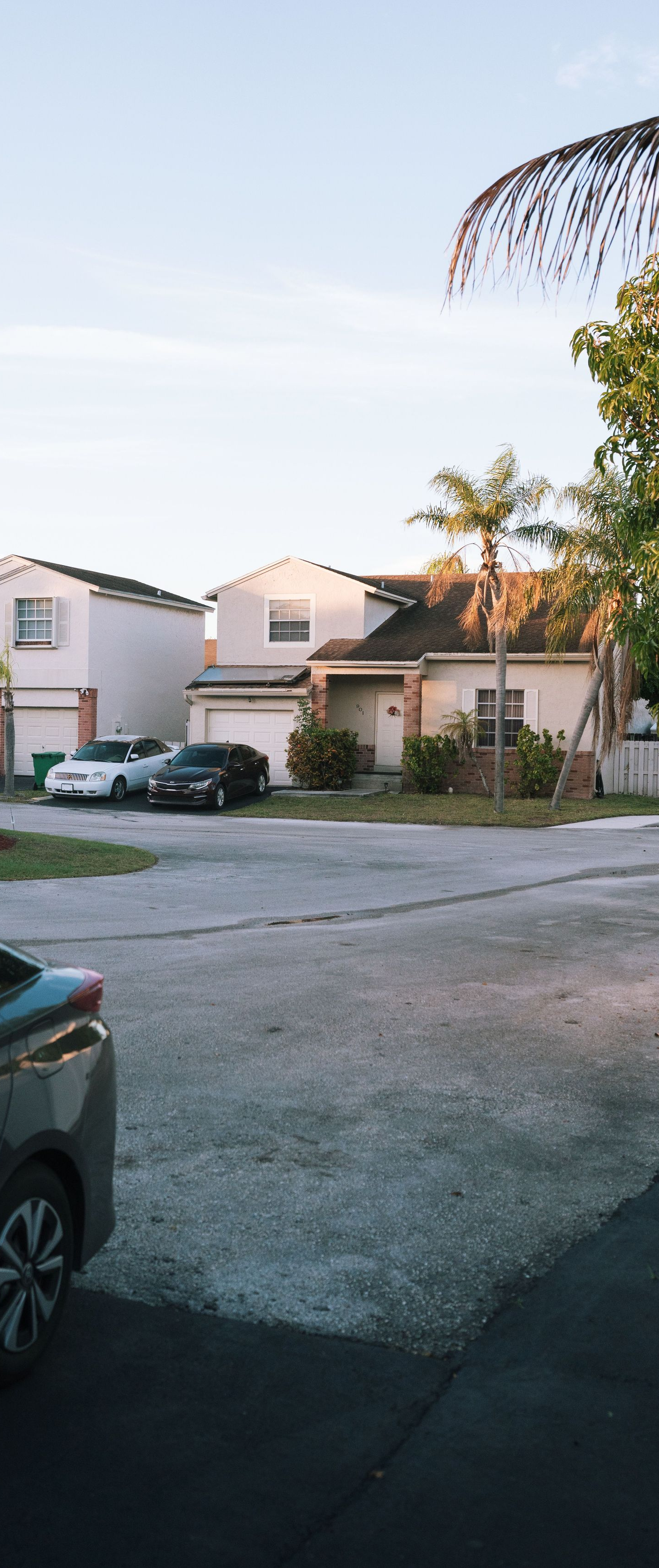 Residential parking lot with two white houses, a parked car, and palm trees at dusk