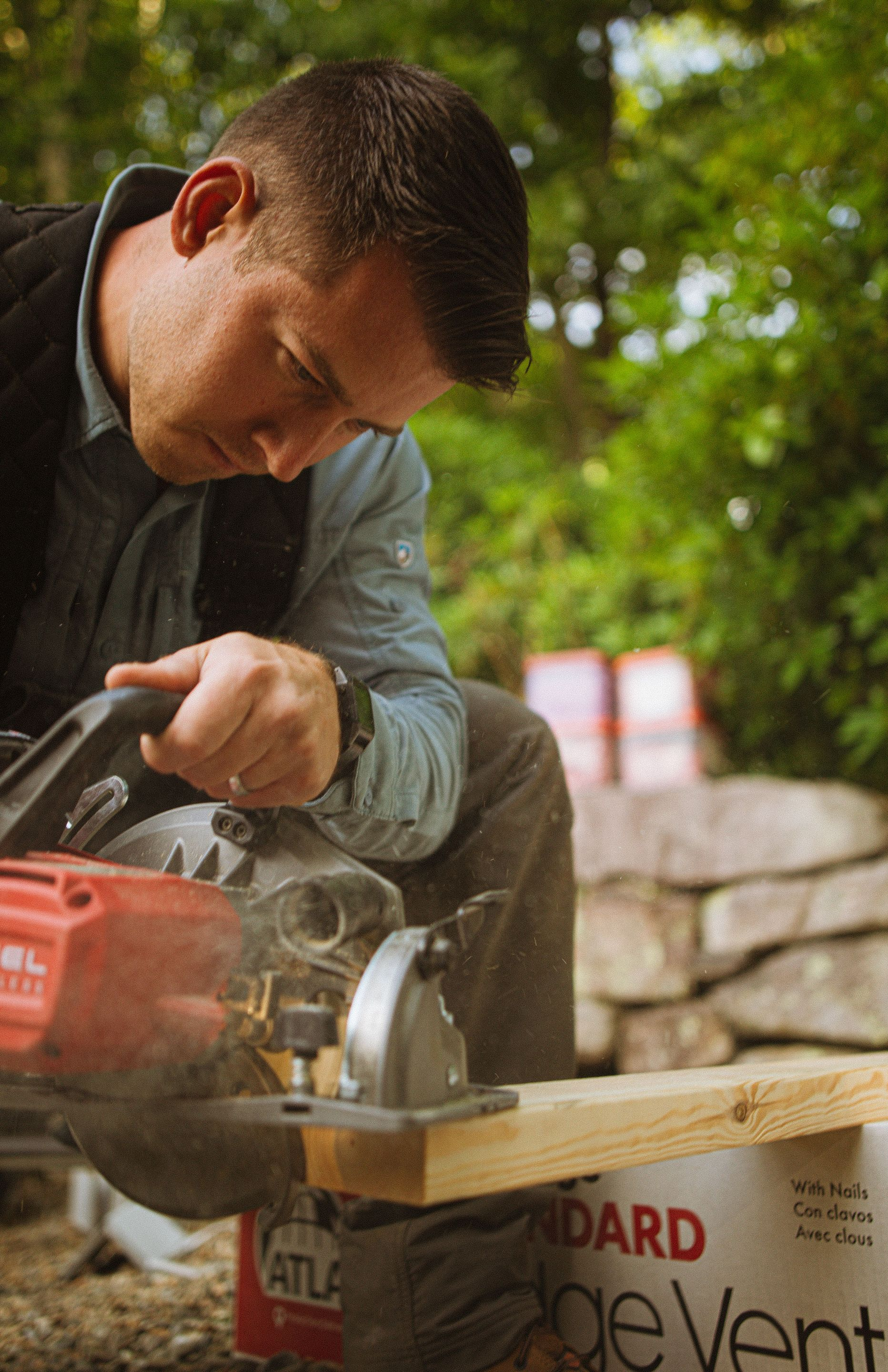 Man using a circular saw to cut a wooden plank outdoors on a stone wall.