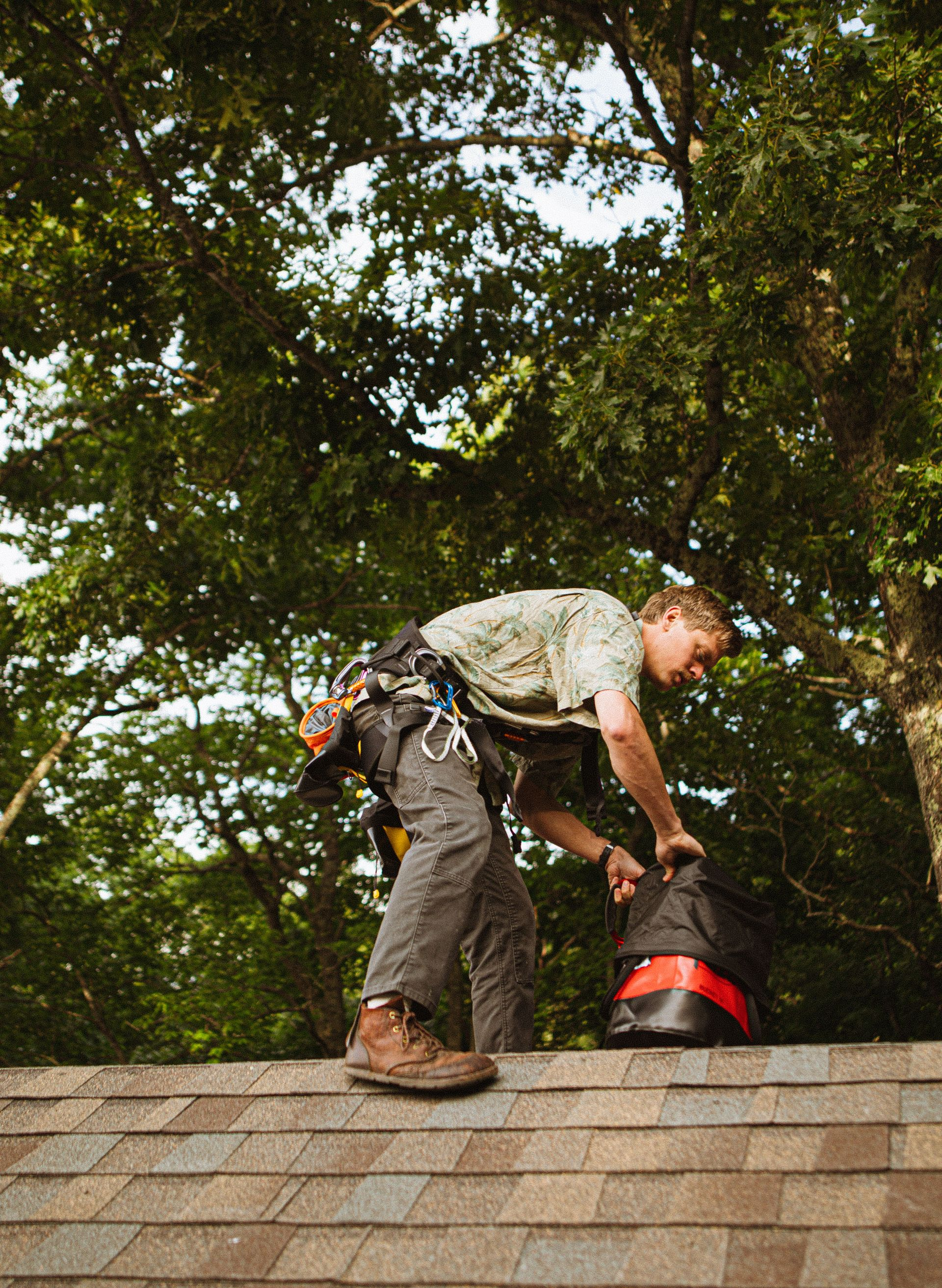 Worker crouches on a roof, handling a red bucket against a backdrop of trees.
