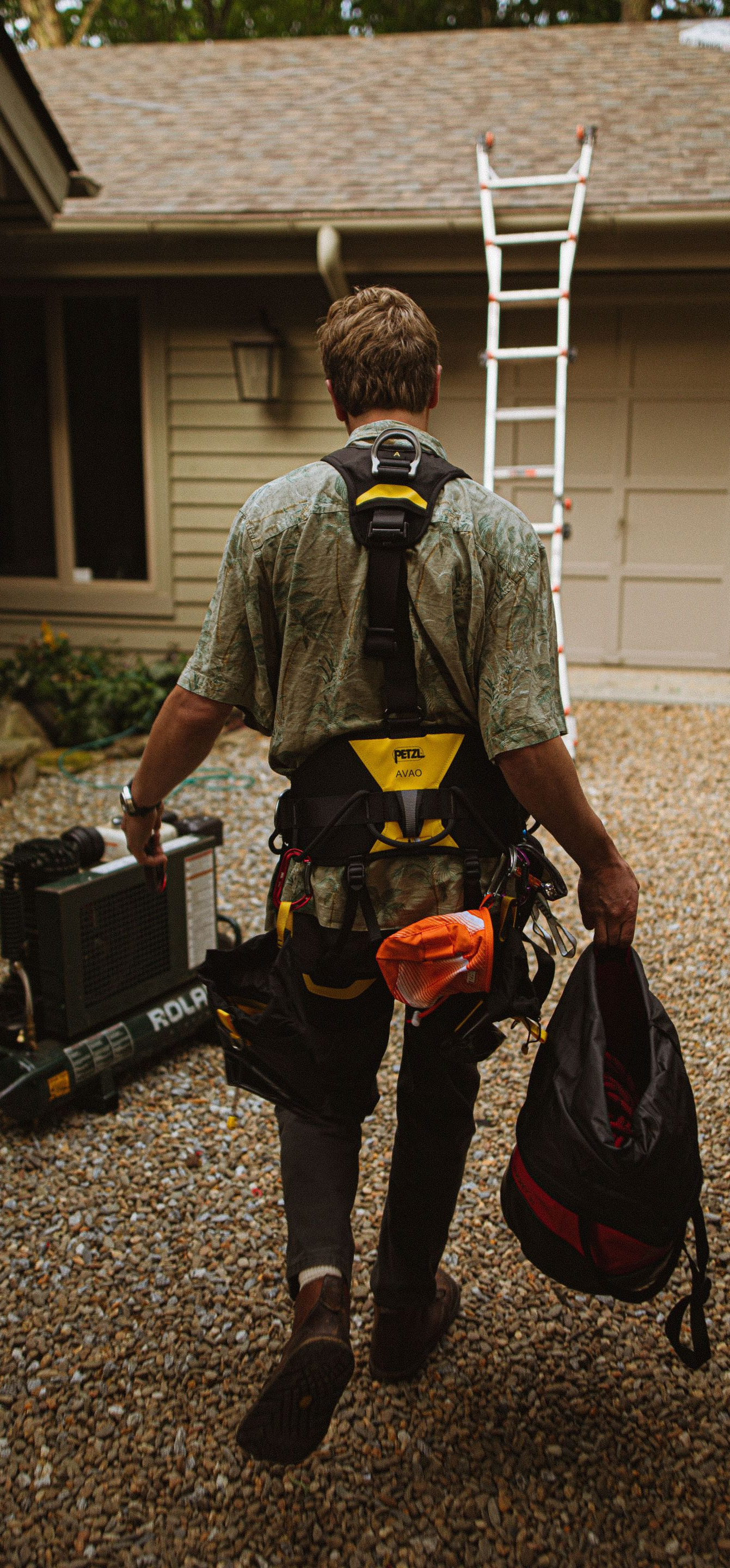 Worker carrying tools toward a house with a ladder leaning against the roof.