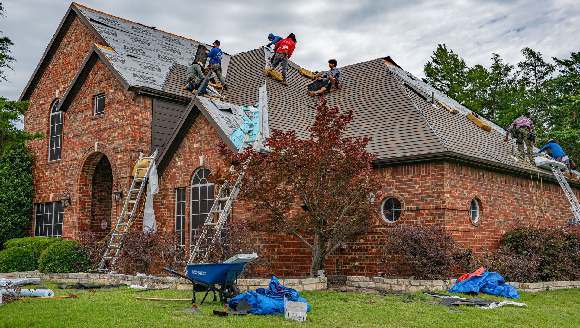 Workers repairing a red brick house roof with blue tarps and wheelbarrow in the yard