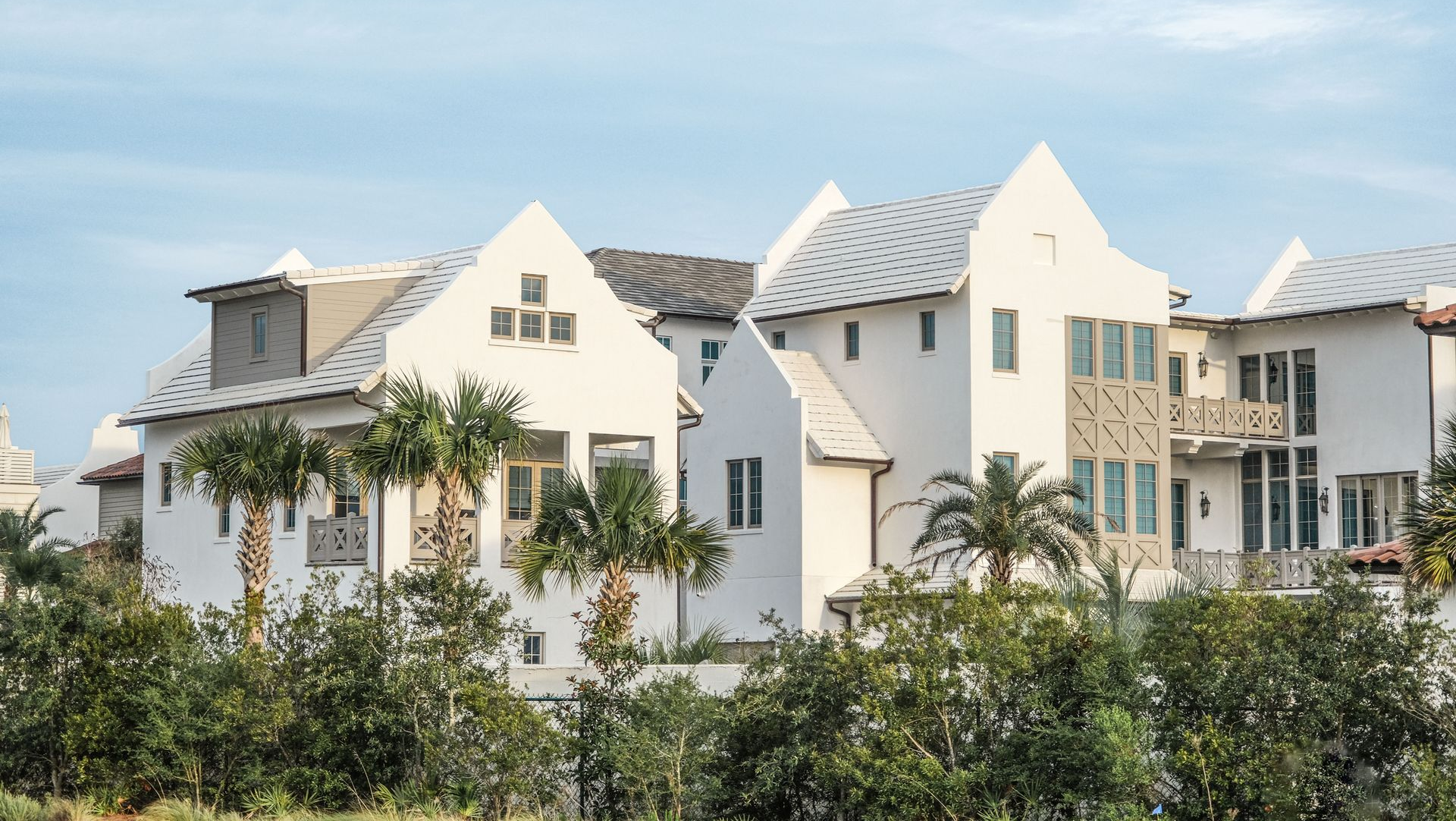Row of white modern houses with peaked roofs behind green shrubs under a bright sky