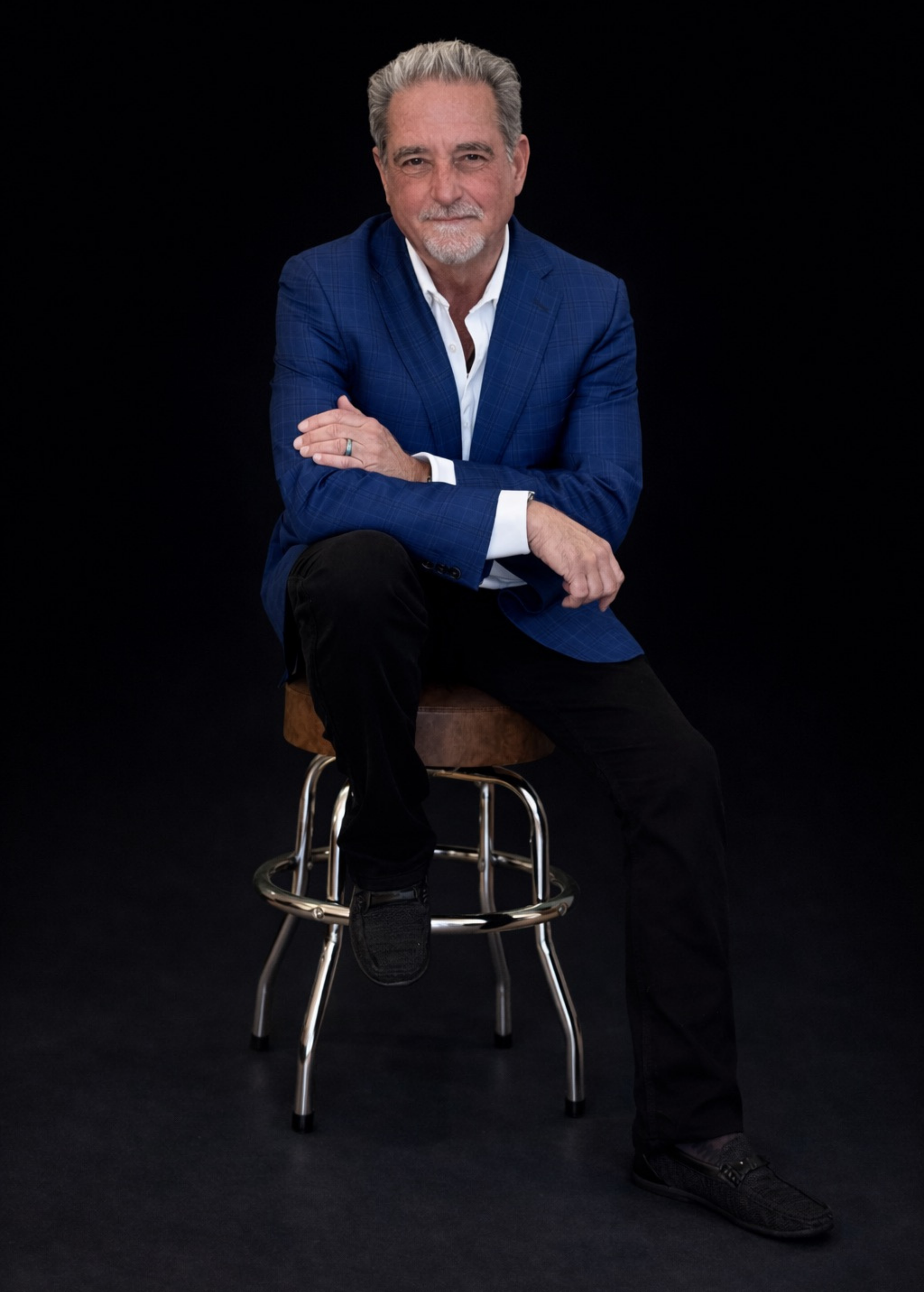 Man in a blue blazer sitting on a stool against a black background.