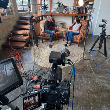 Two people talking on a couch in a studio, with cameras and lighting equipment set up around them.