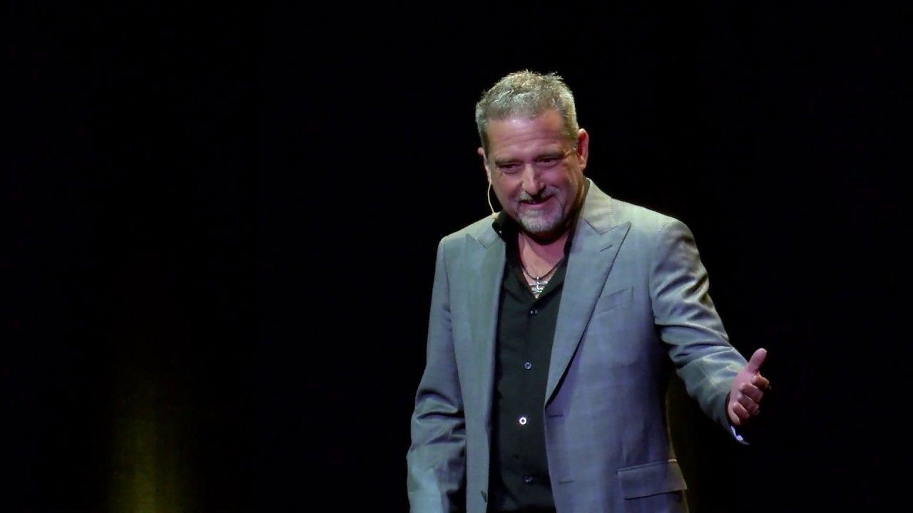 A man in a suit and black shirt is giving a speech on a stage.