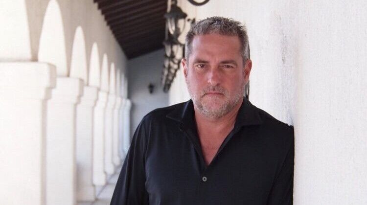 A man in a black shirt leans against a white wall