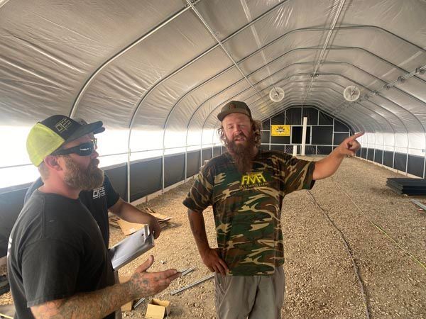 Two men standing inside a long white arched greenhouse or tunnel, one pointing ahead.