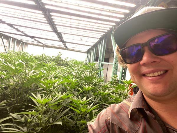 Person smiling in sunglasses beside lush green plants under a greenhouse roof