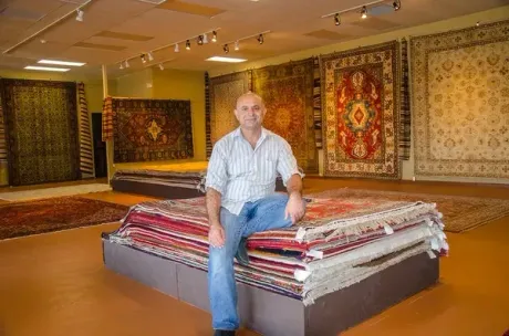 A man is sitting on a stack of rugs in a rug store.