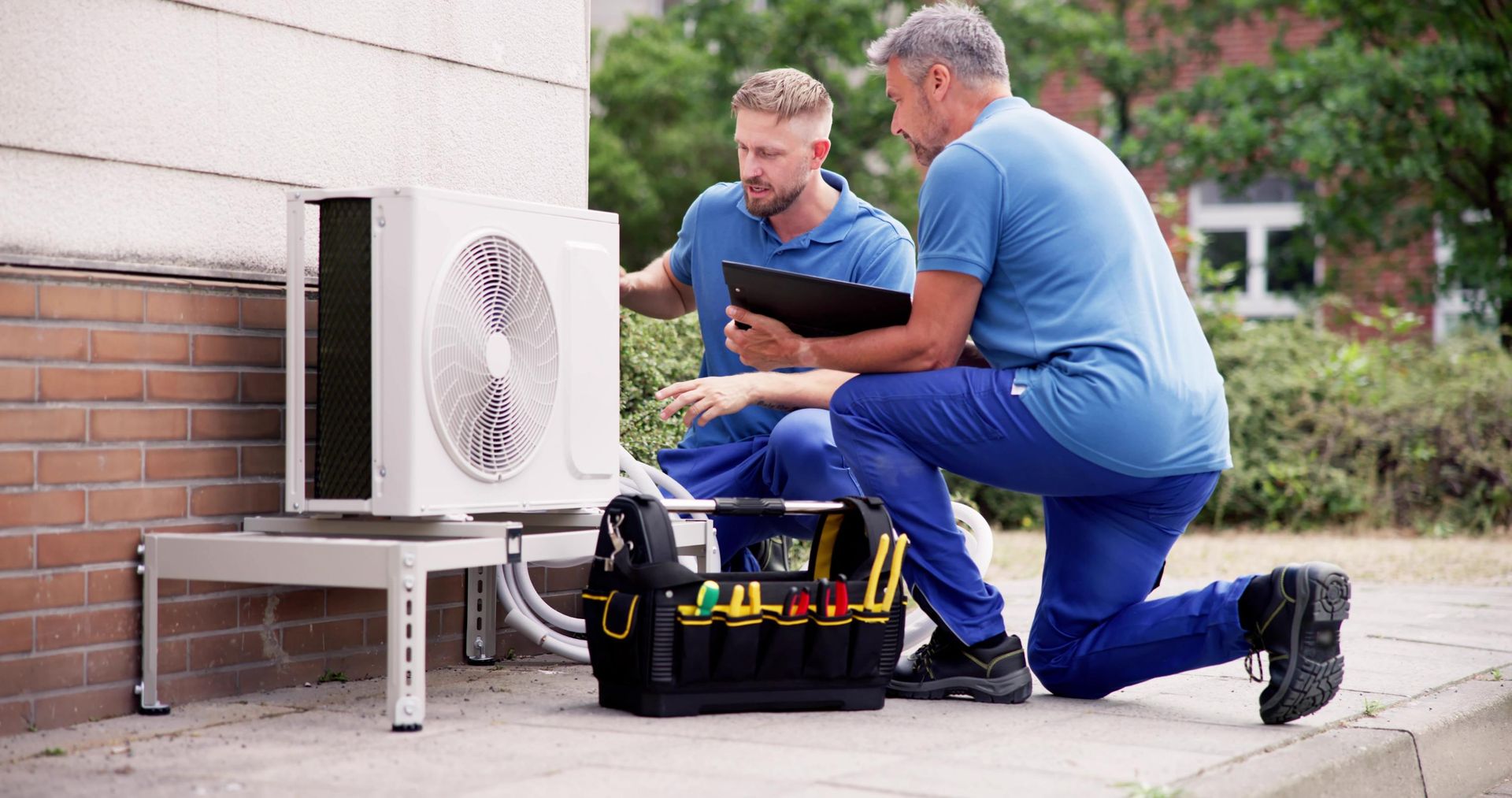 Two men are working on an air conditioner outside of a building.